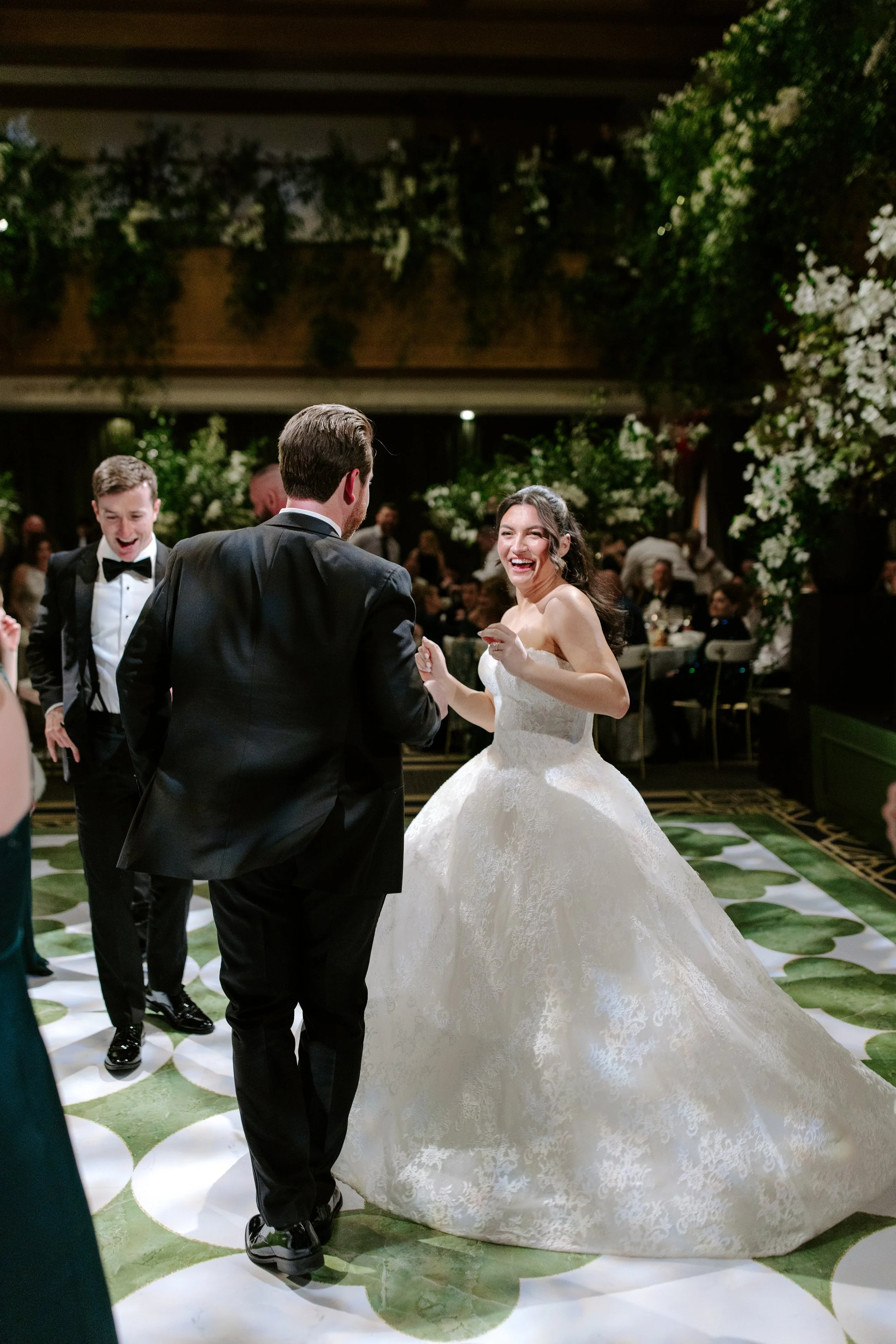 A bride in a white wedding gown dancing with a groom in a black tuxedo at a wedding reception, surrounded by guests.