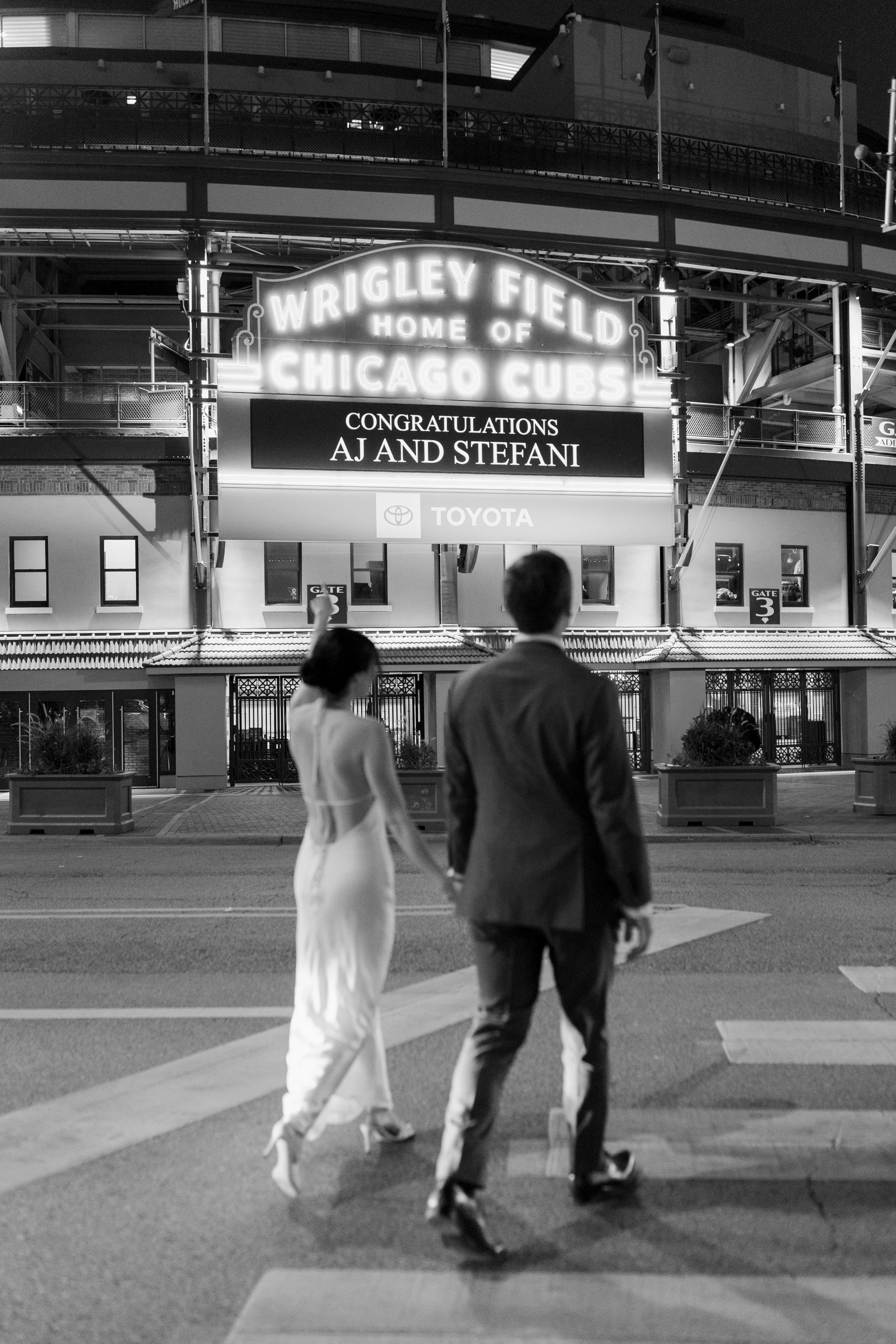 A black and white photo of a man and woman crossing a street at night with a stadium in the background. The stadium marquee reads 'Wrigley Field, Home of Chicago Cubs' and congratulates AJ and Stefani.