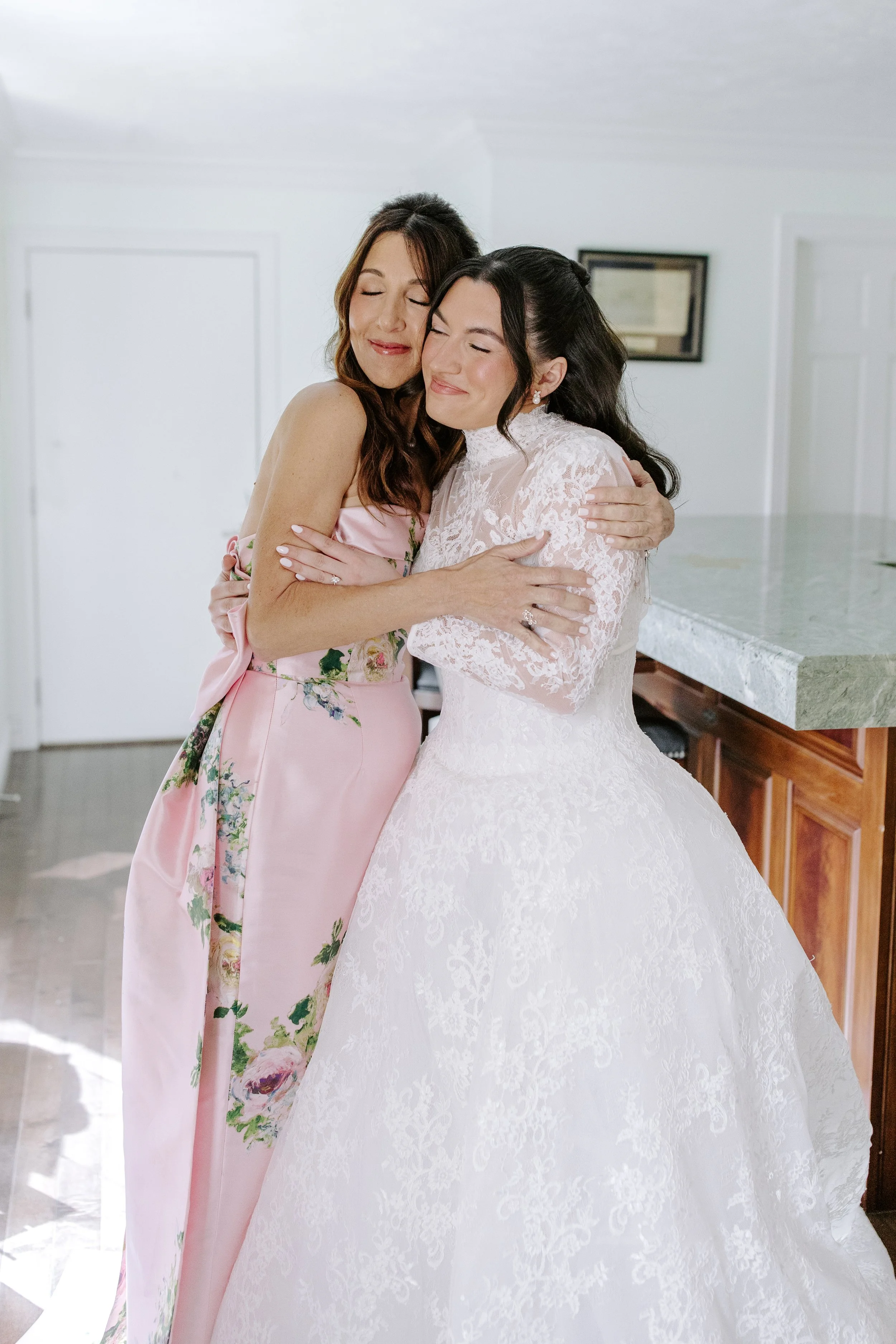 Two women hugging, one in a pink floral dress and the other in a white lace wedding gown, in a bright indoor setting.