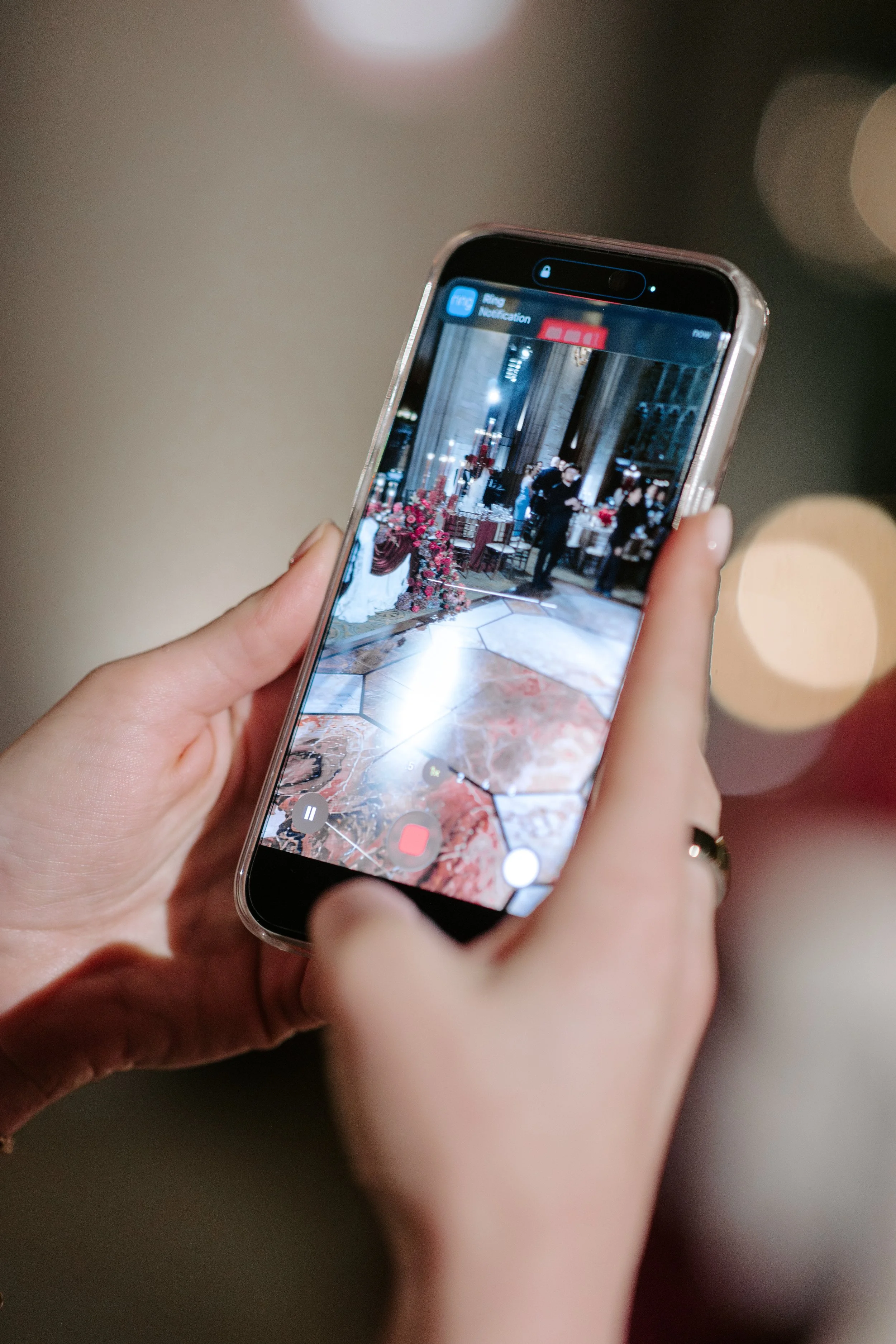 Close-up of a person holding a smartphone, recording or taking a photo of an indoor event with people and floral arrangements.