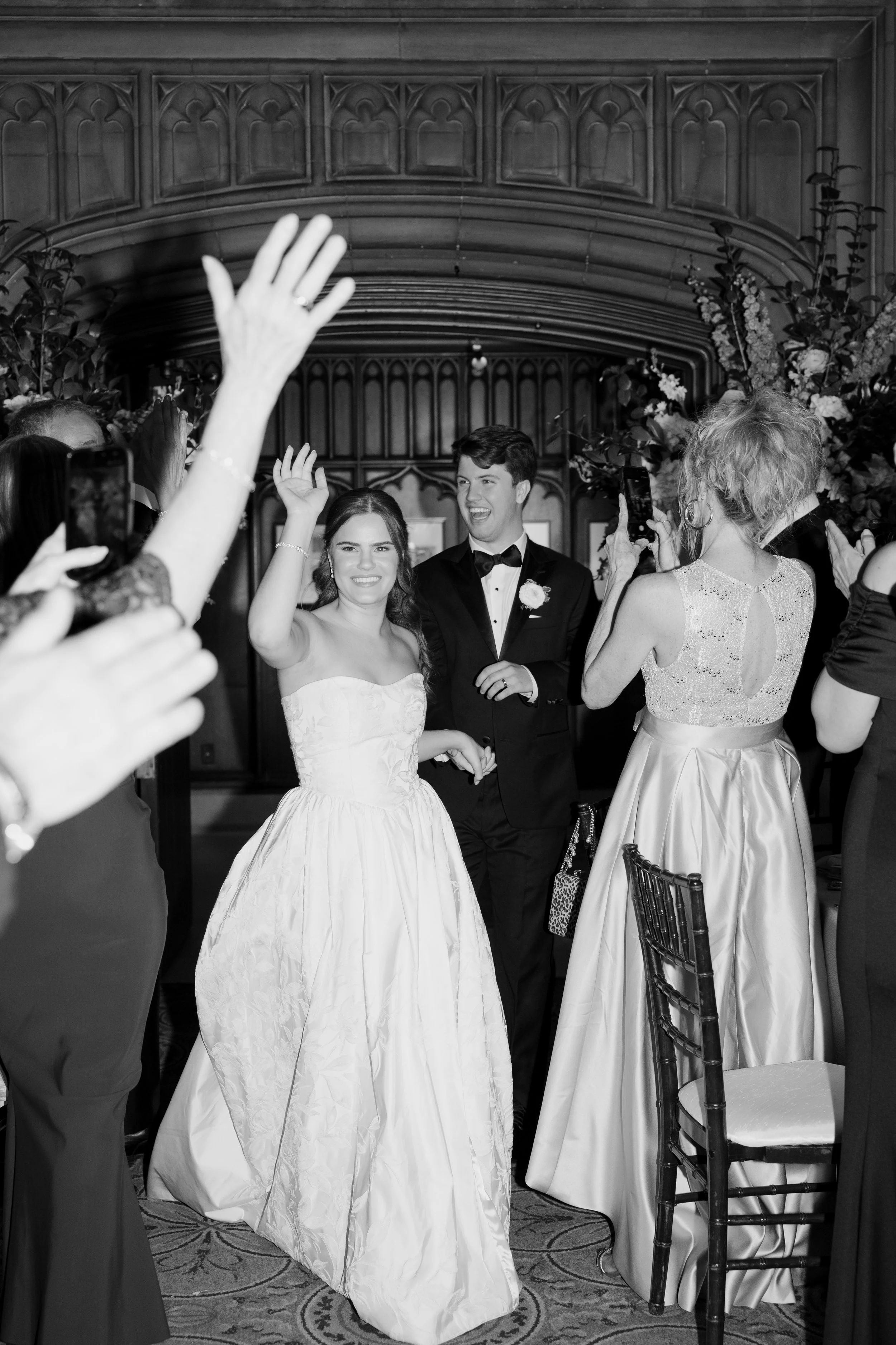 A bride and groom are smiling and waving at a wedding reception. The bride is wearing a strapless white wedding gown and the groom is dressed in a tuxedo with a bow tie. They are surrounded by guests taking photos and clapping, and the background fea
