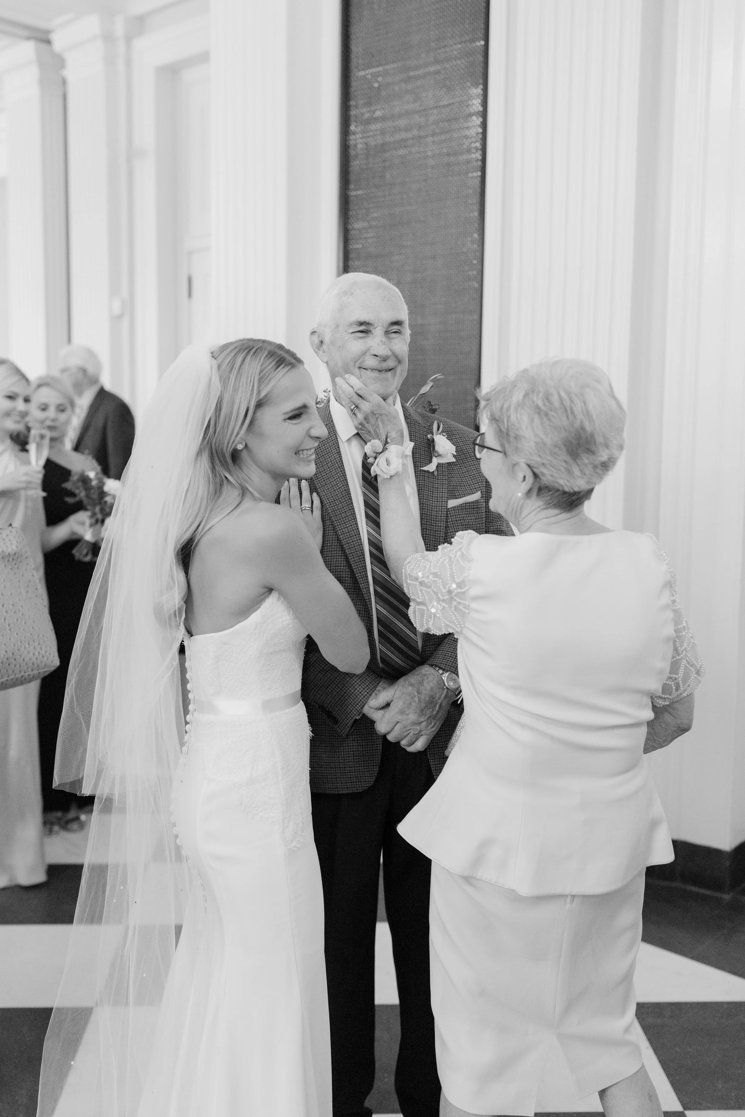 A bride in a wedding dress and veil, smiling with her hand on her face, looks at an elderly woman in a white dress, who is touching her chin. Standing between them is an older man in a checkered jacket with a boutonniere, smiling. In the background, 