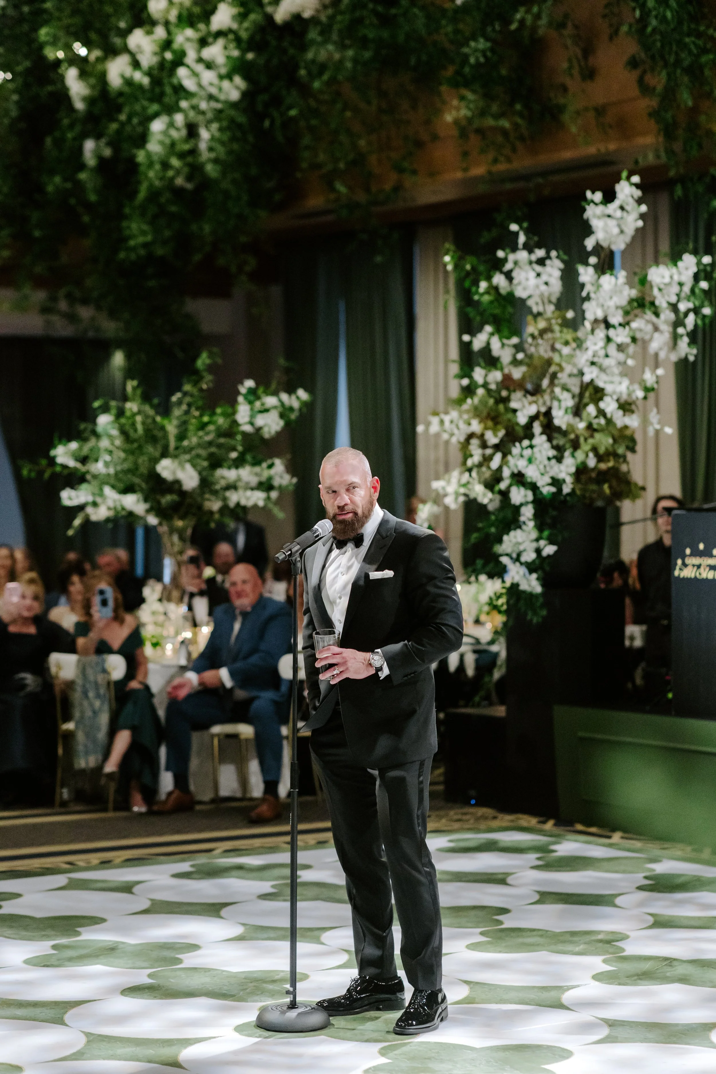 A man in a tuxedo speaking at a formal event with floral decorations and seated guests in the background.