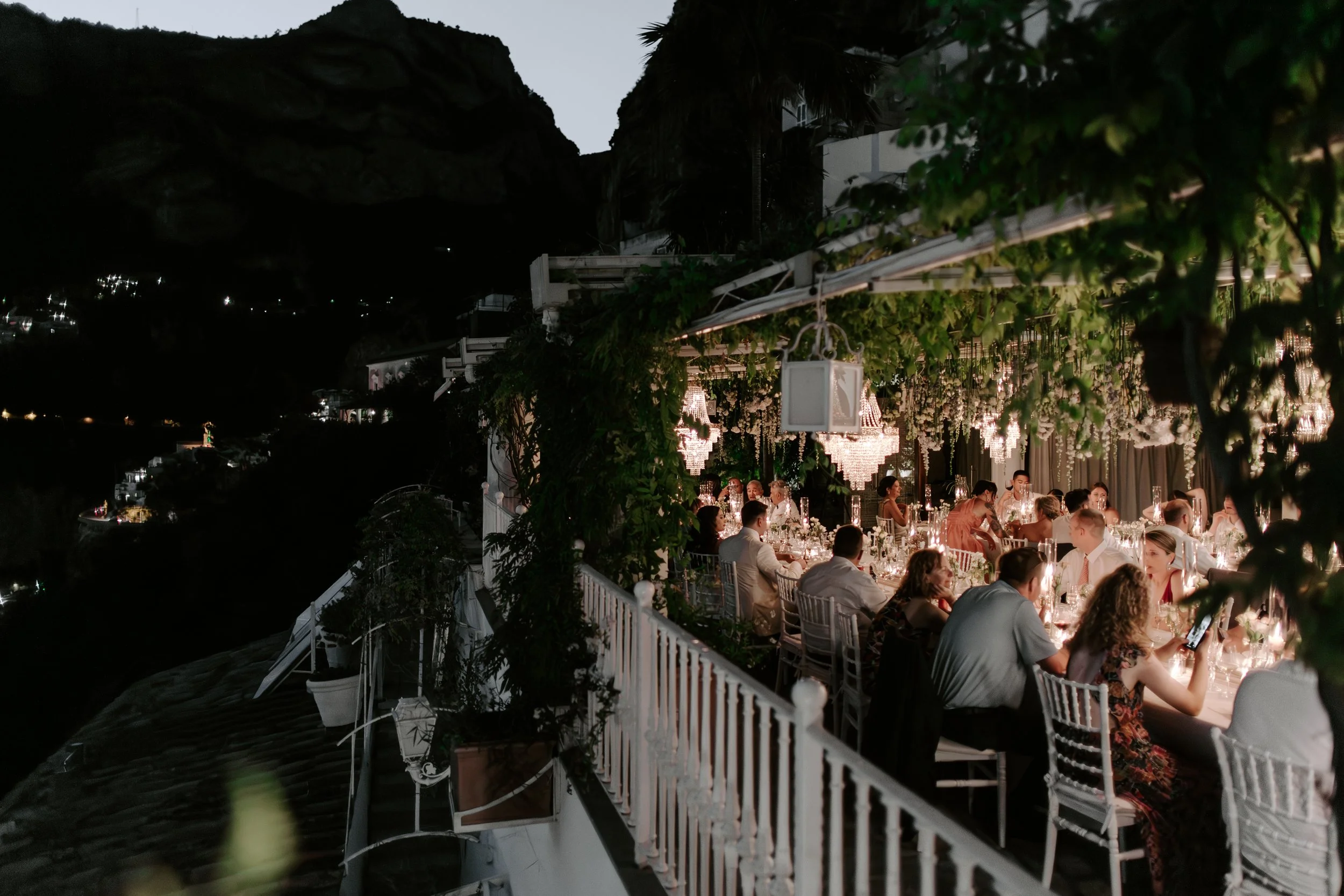 Nighttime outdoor wedding reception with guests sitting at decorated tables, hanging chandeliers, and lush greenery, overlooking a hillside.