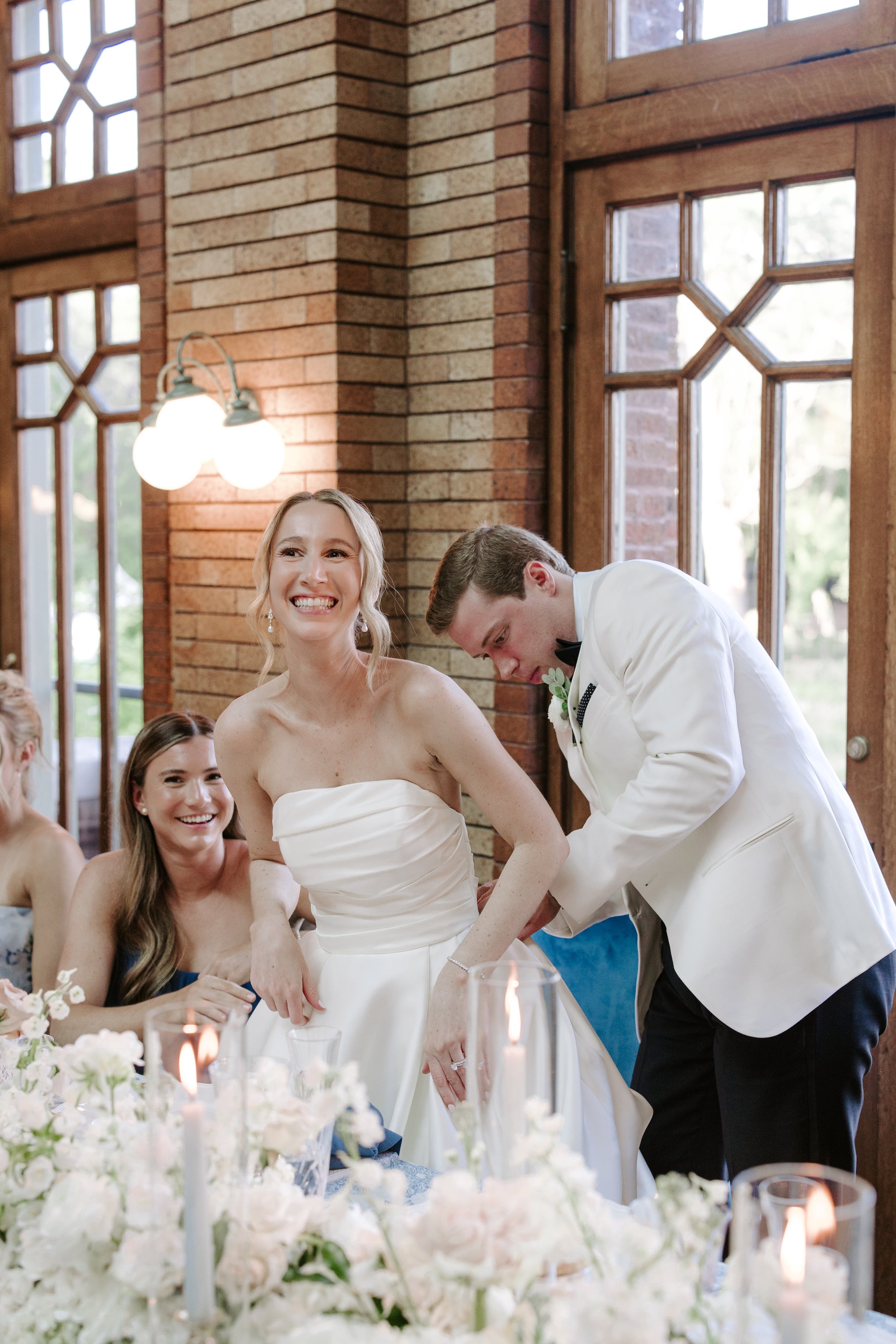 A wedding reception with a bride smiling and a groom in a white tuxedo leaning over to help the bride with her dress, surrounded by bridesmaids and floral decorations.