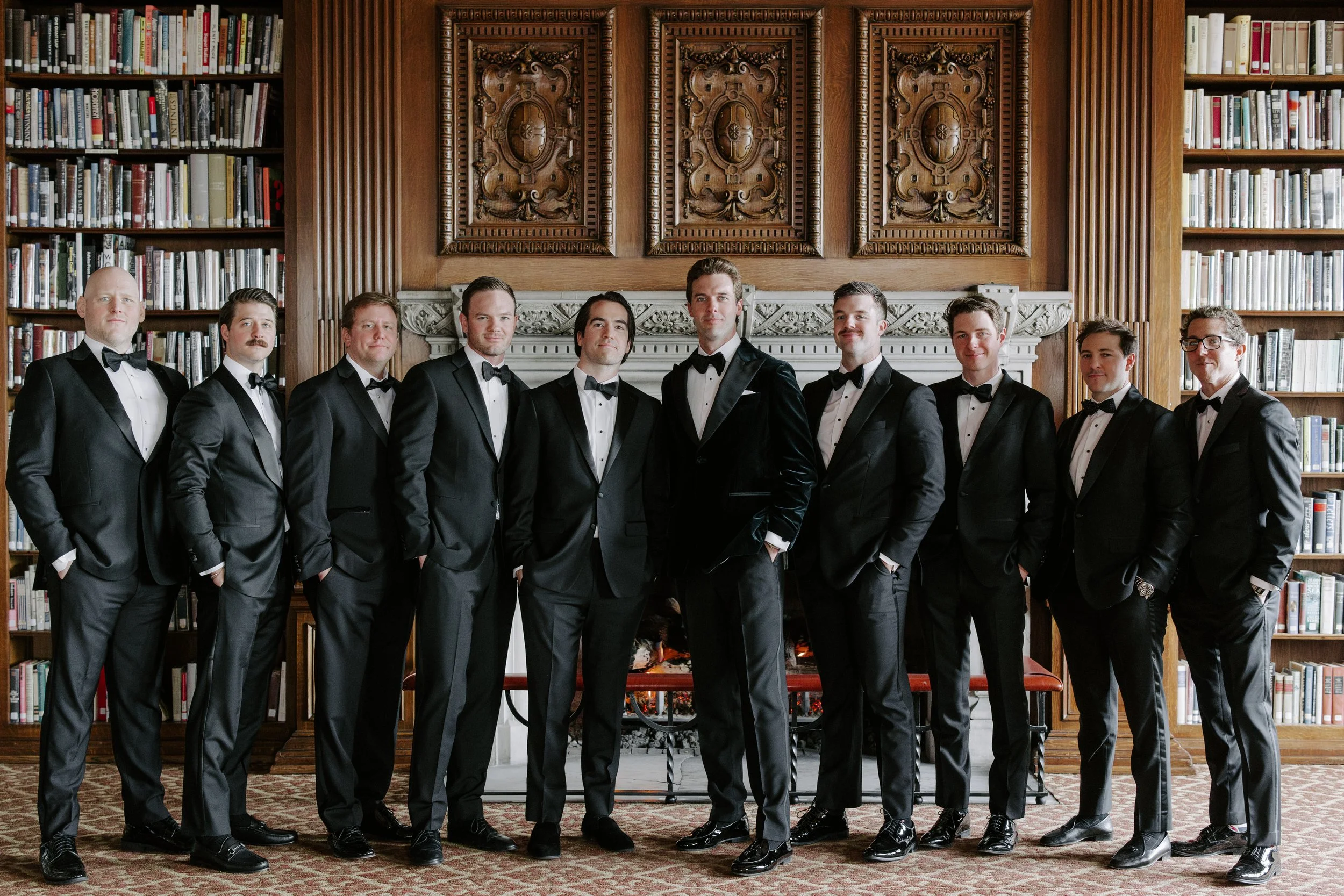 A group of eleven men dressed in black tuxedos with bow ties standing in a line in front of a fireplace and bookshelves in a library or formal room.
