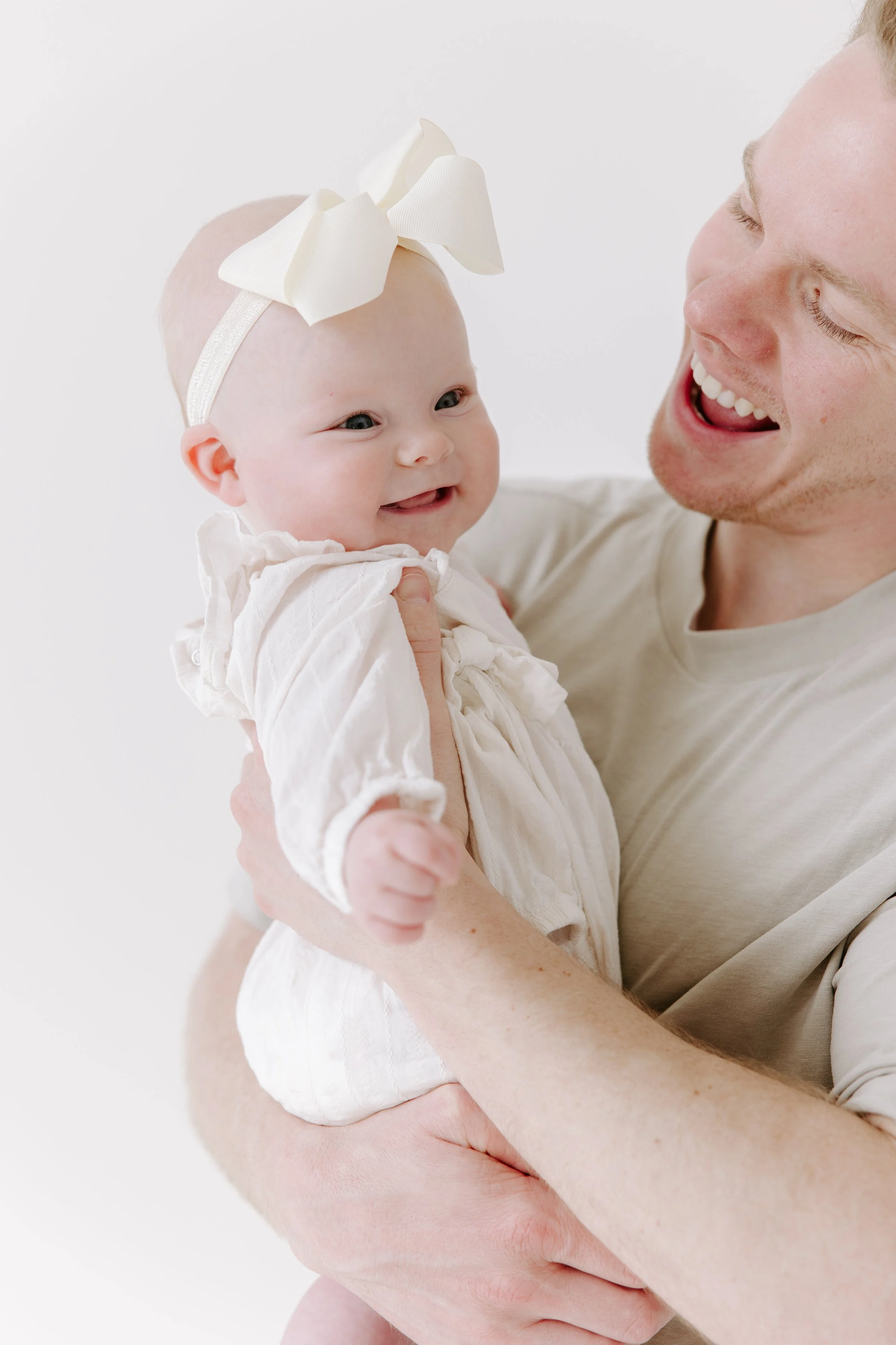 A man with light skin and short blond hair holding a smiling baby girl with a large white bow headband and a white dress, both facing the camera, against a plain white background.