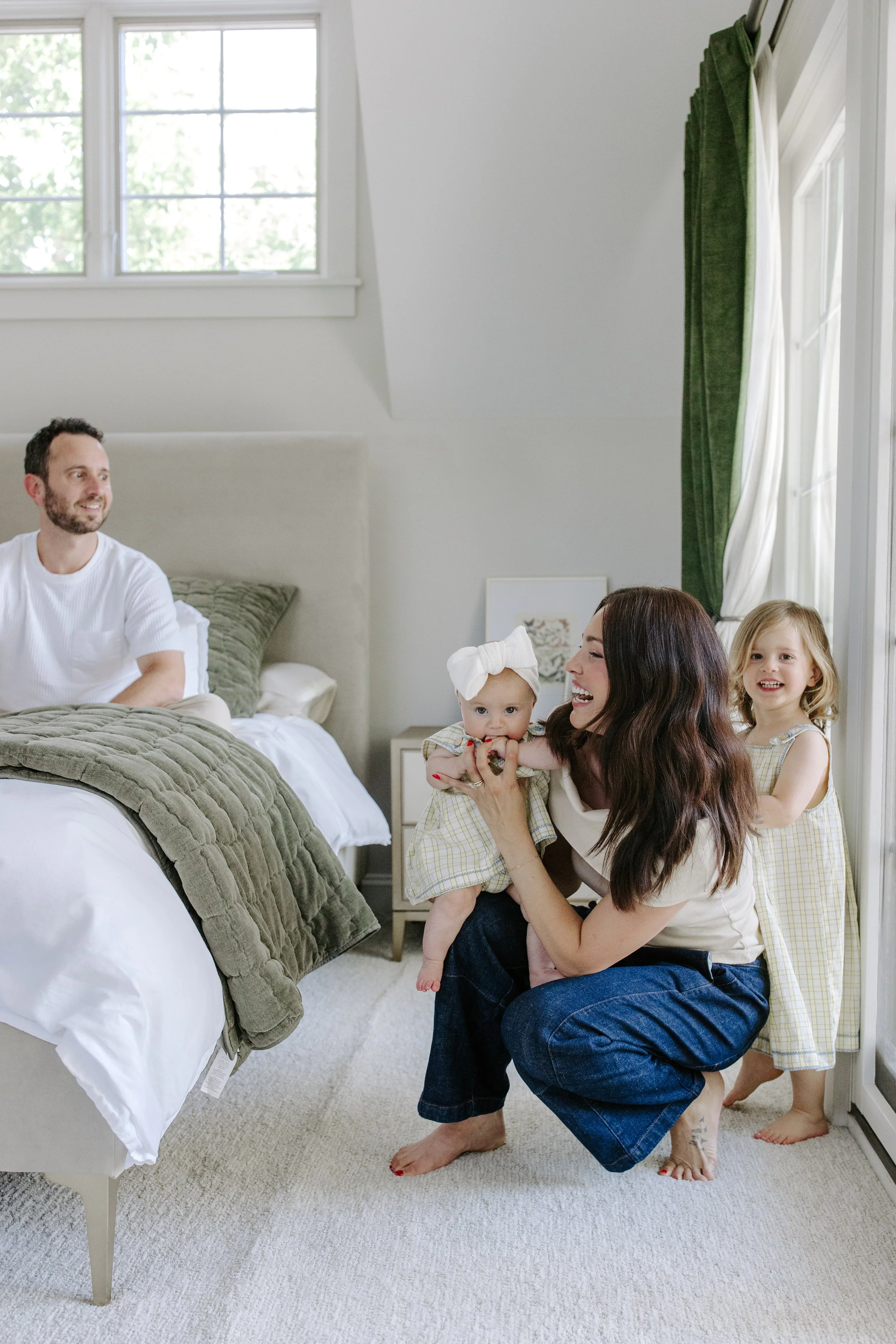 A family with young children in a bedroom, with two adults and two children, one of whom is holding a baby, all smiling and interacting.
