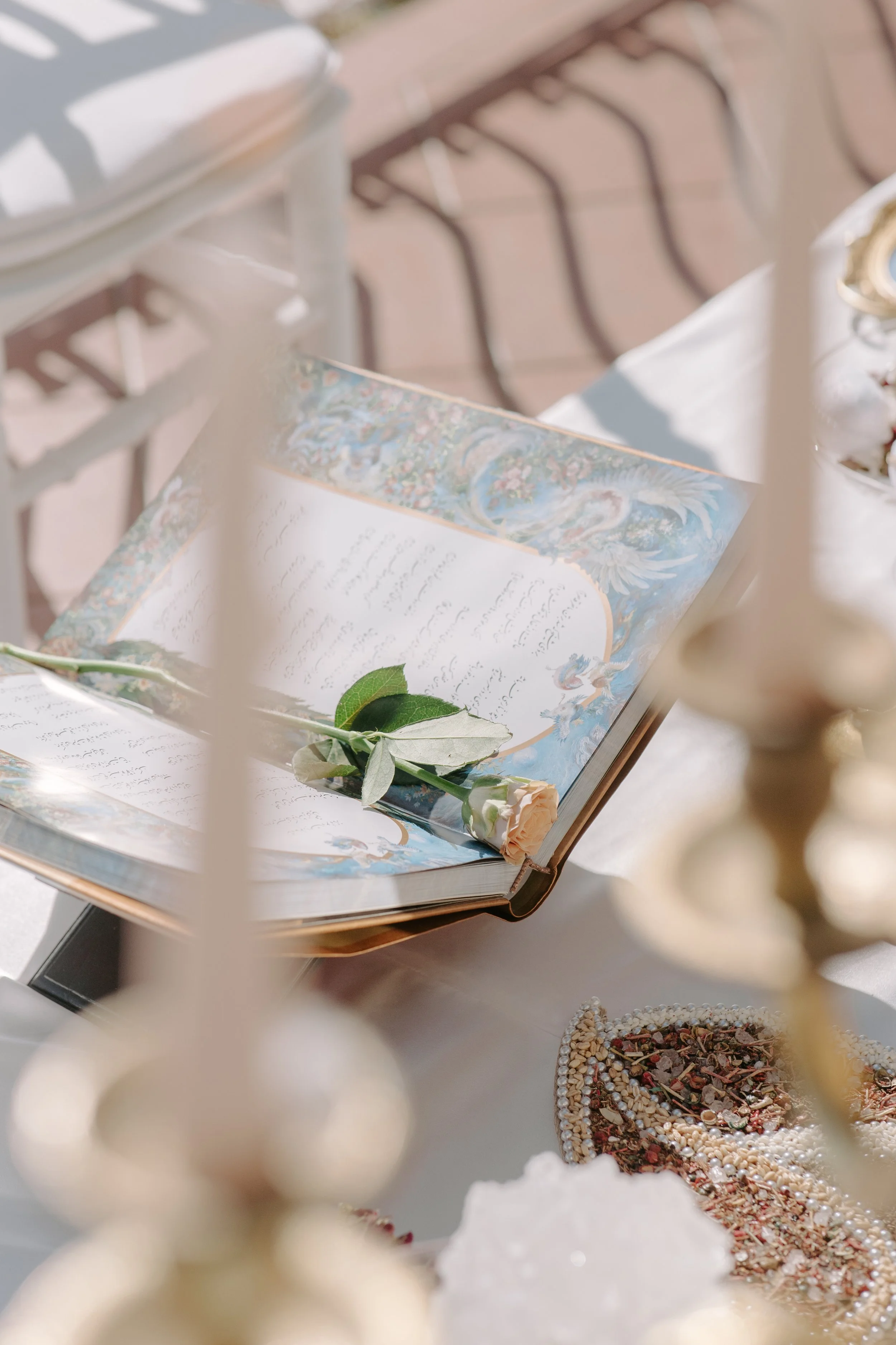 Open book with illustrated cover featuring a peacock and floral design, on a table next to a single peach-colored rose, surrounded by decorative items including a beaded tray with dried herbs and a glass container. Out-of-focus chairs are in the back