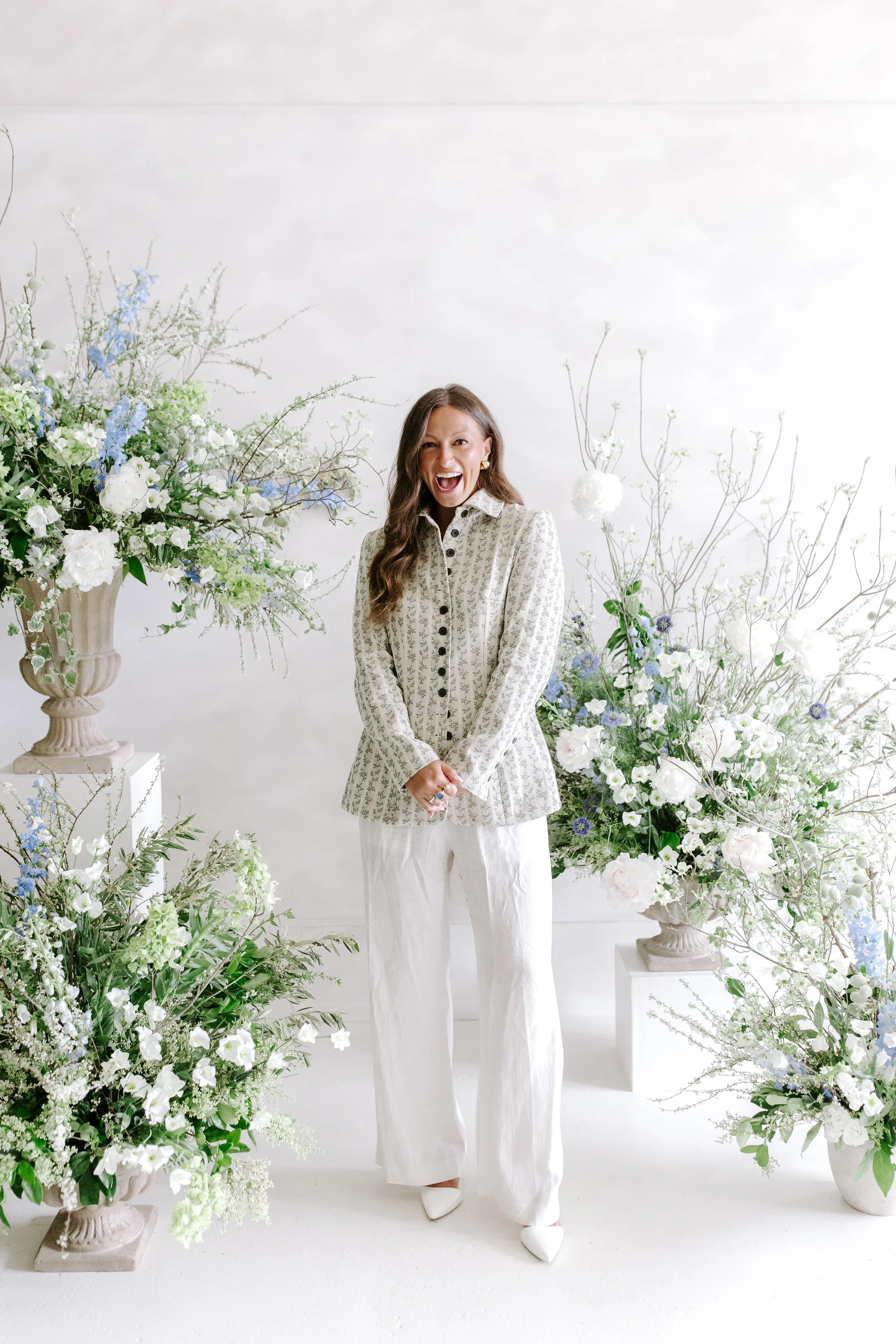 A woman laughing in a white floral blouse and white pants, standing among white and pastel-colored floral arrangements in a minimalist room.