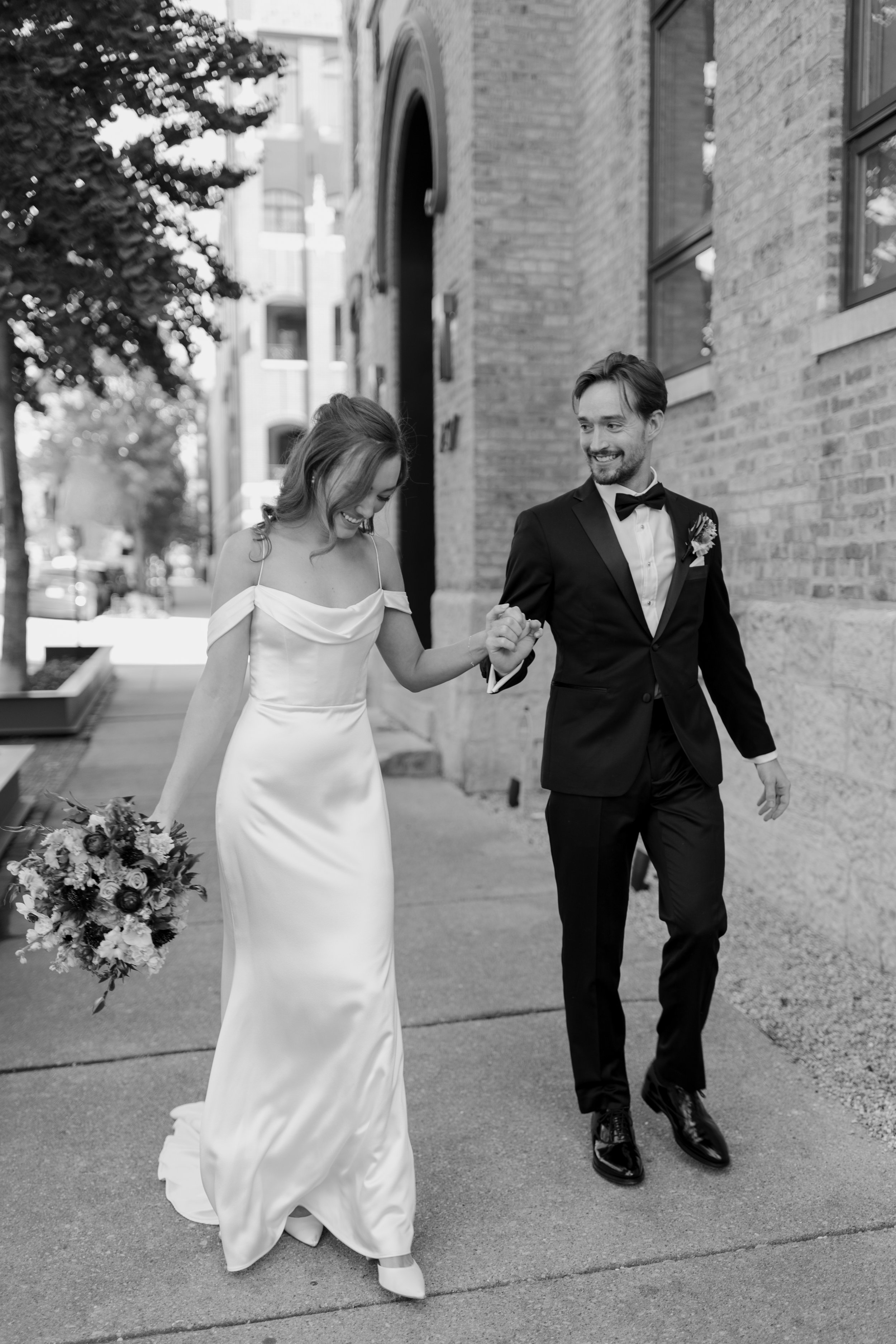A bride and groom walking hand in hand outside a building. The bride is wearing a white wedding dress and holding a bouquet of flowers. The groom is dressed in a tuxedo with a bow tie. They are smiling and appear happy.