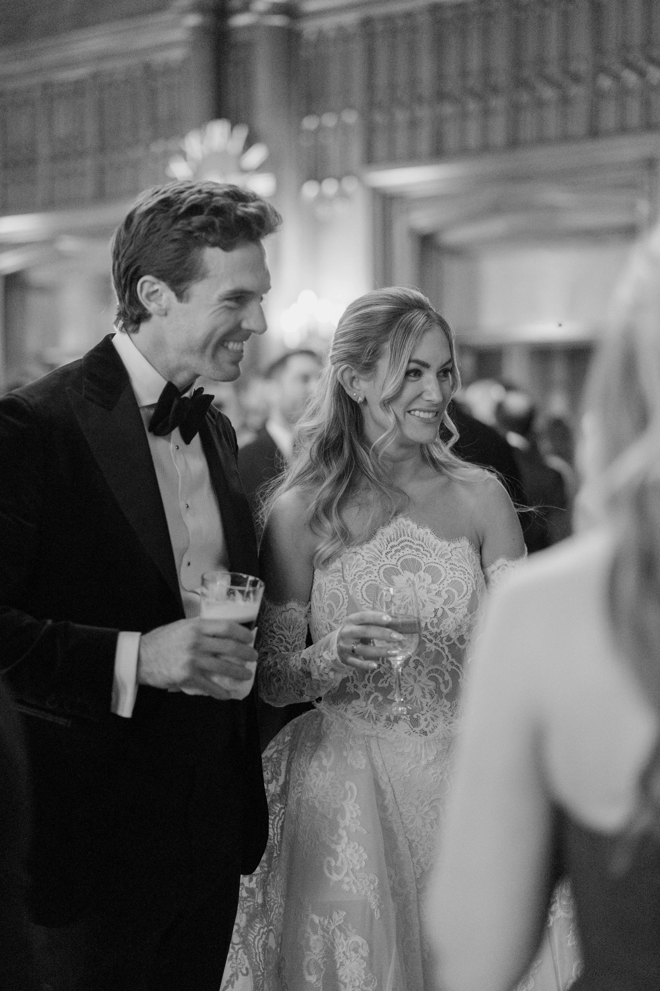 A black and white photo of a man and woman at a formal event, holding drinks, smiling, dressed in formal attire with the man in a tuxedo and the woman in a lace dress.