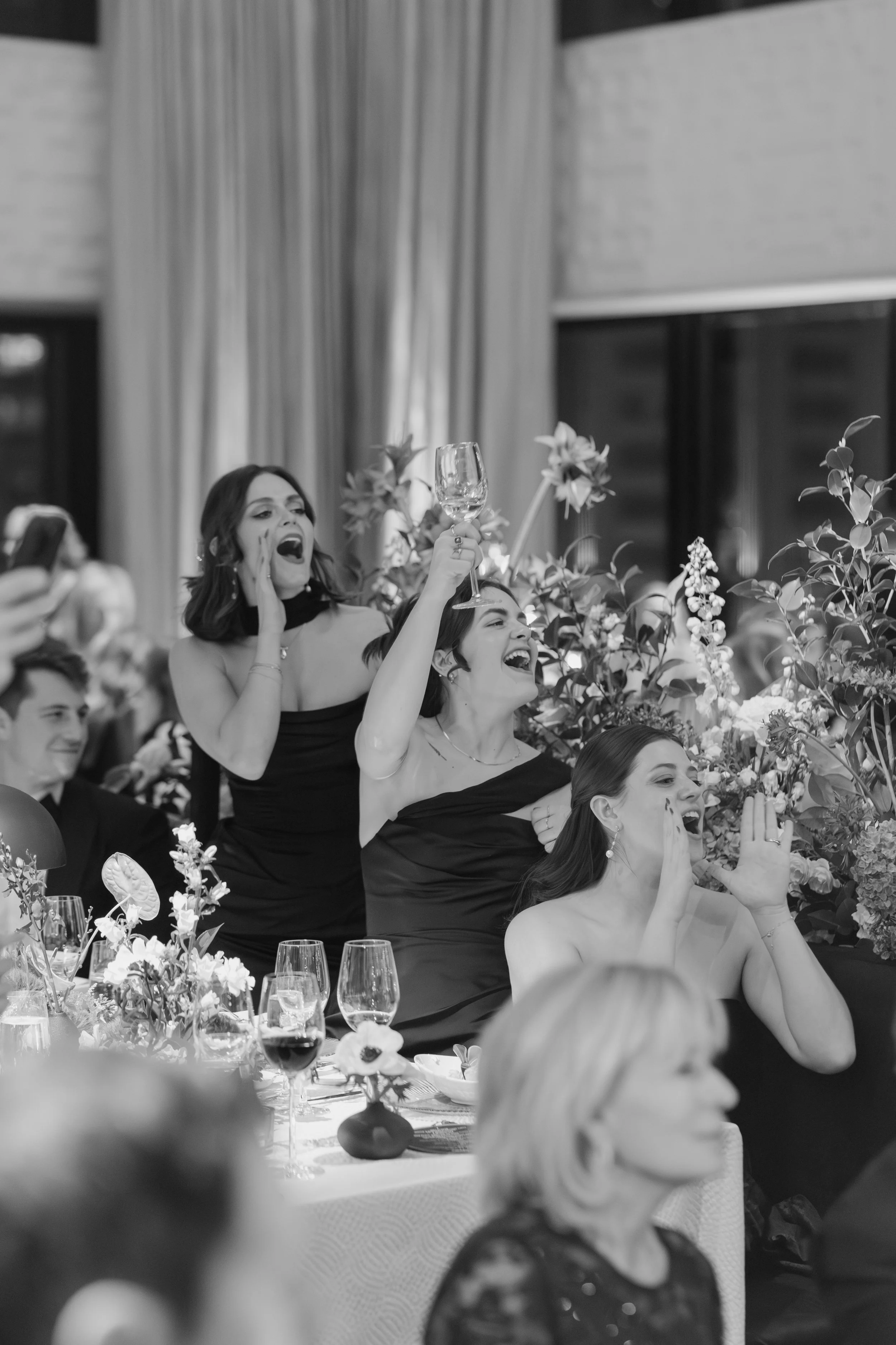 People celebrating at a formal event with flowers, wine glasses, and decorated tables, with some individuals raising glasses and cheering.