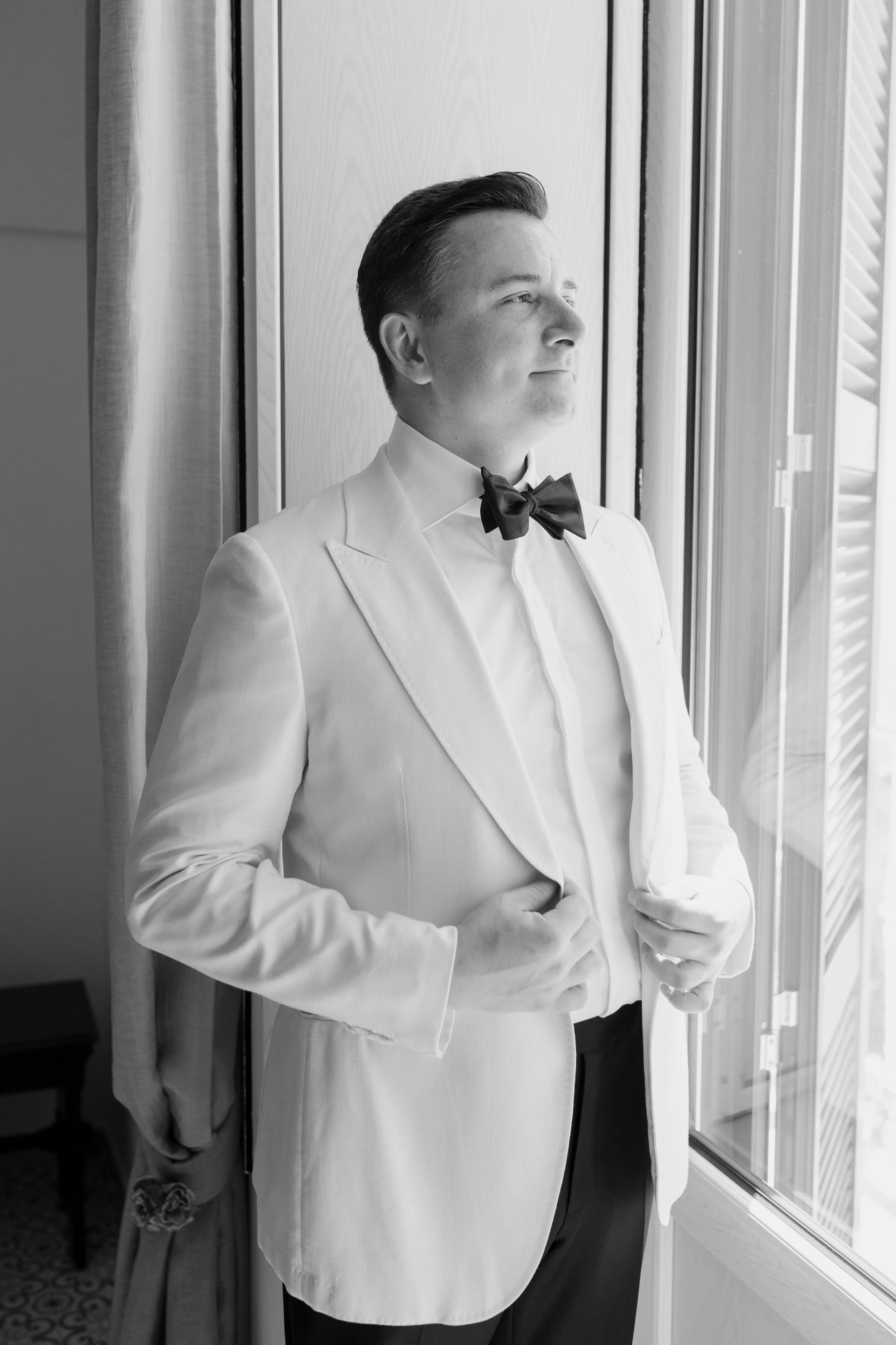 A man dressed in a white tuxedo with a black bow tie standing indoors near a window, looking outside with a confident expression.