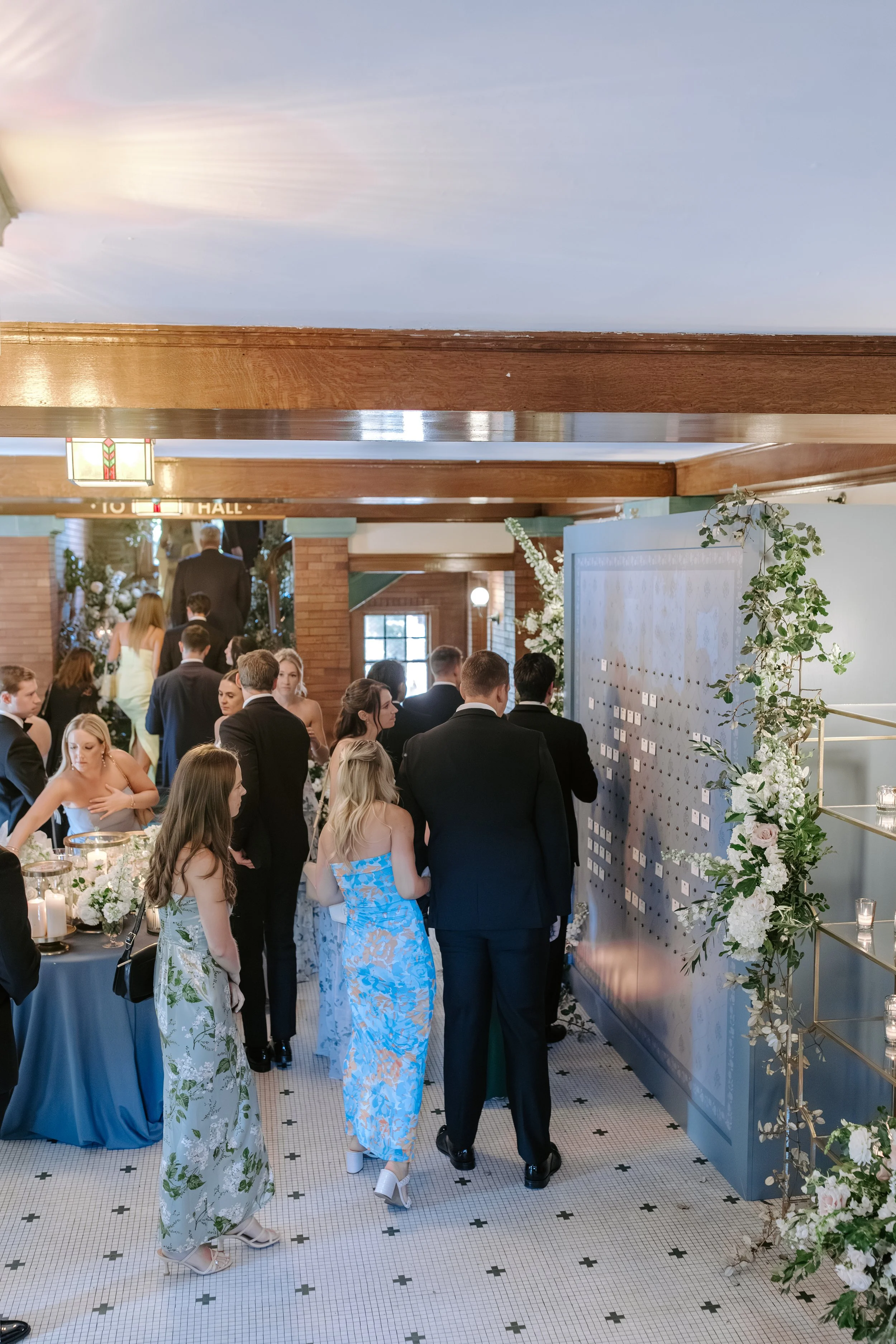 People dressed in formal attire at a wedding reception, standing and talking near floral arrangements and candles, with a wooden beam and decorative wall in the background.