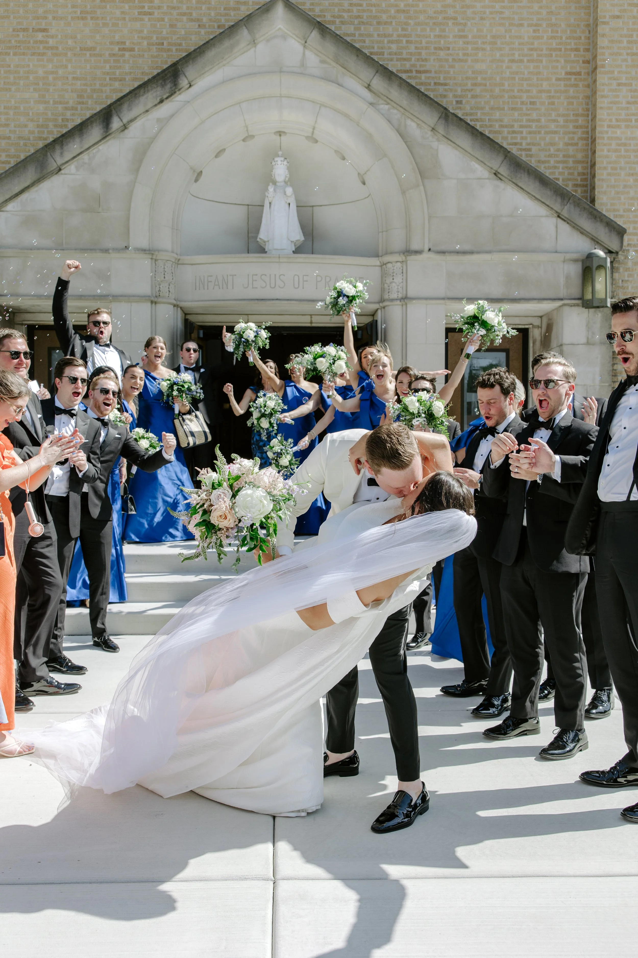 A bride and groom kiss in front of a crowd of people celebrating outside a church. The guests are clapping, cheering, and holding bouquets. The church has a sign reading 'Infant Jesus of Pra' and a statue of the infant Jesus above the entrance.