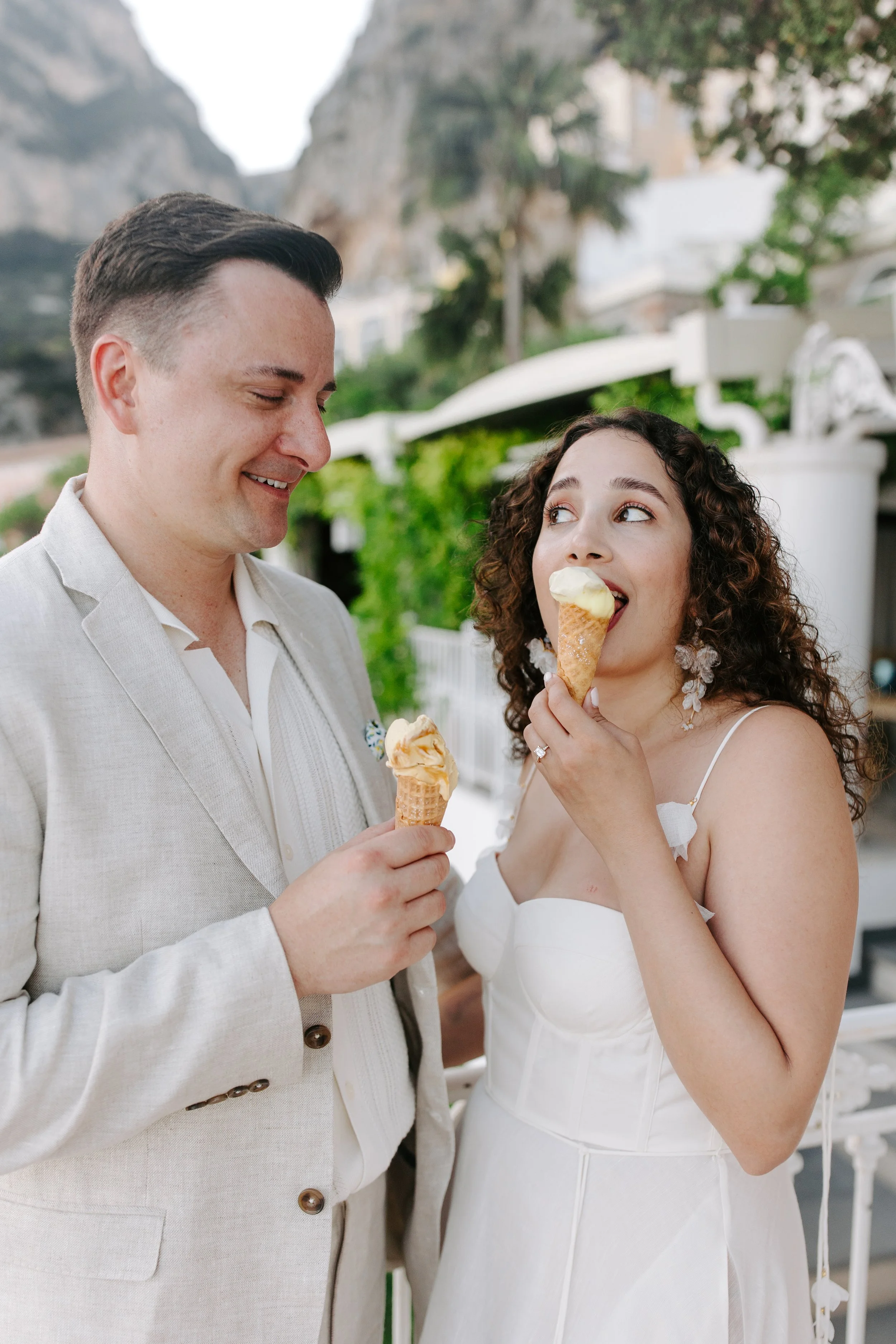 A couple dressed in wedding attire sharing ice cream cones outdoors with rocky hills and greenery in the background.