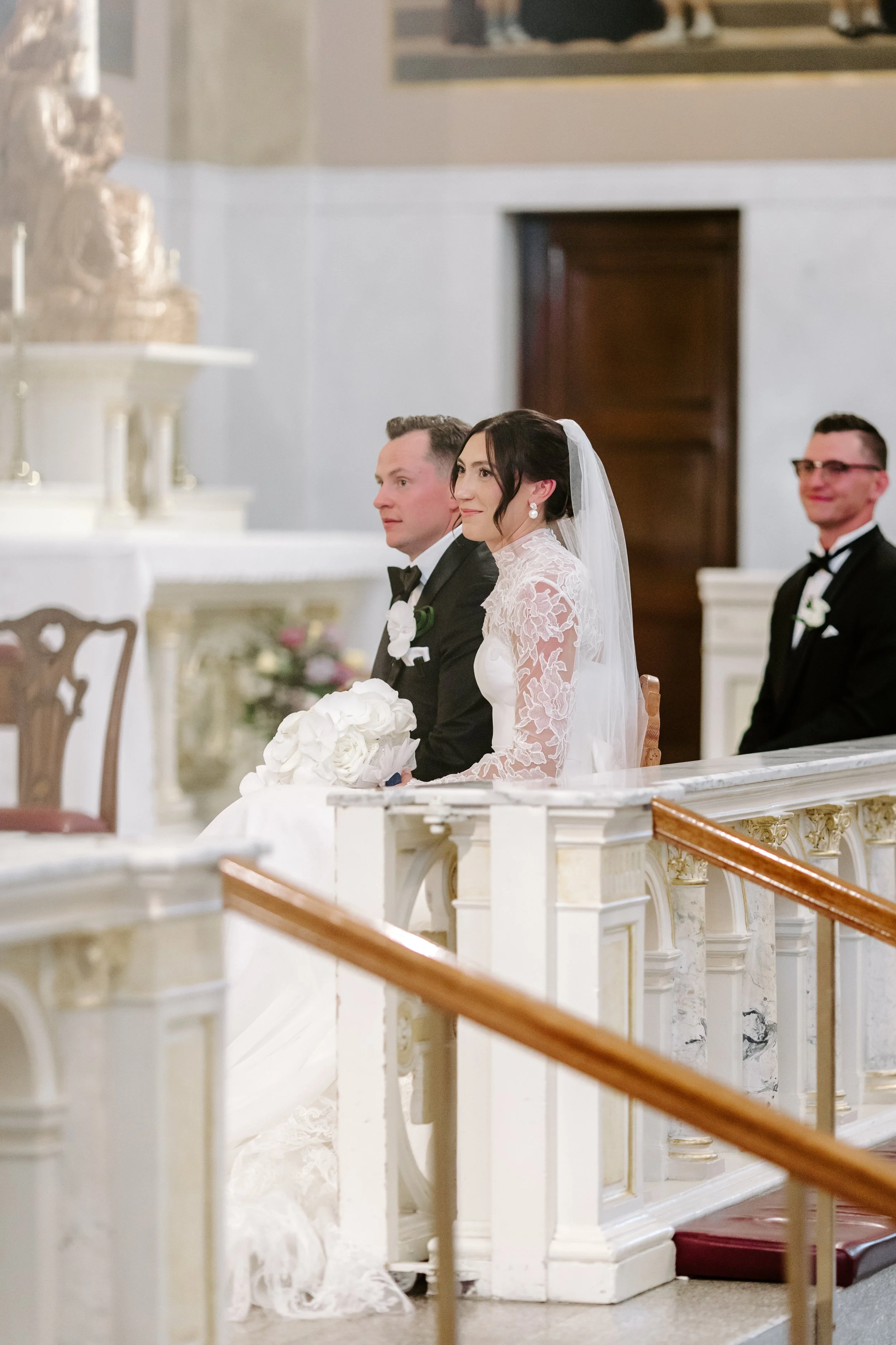 A bride and groom seated during a wedding ceremony inside a church, with the bride in a lace wedding dress and veil, and the groom in a tuxedo, along with another man in a tuxedo.