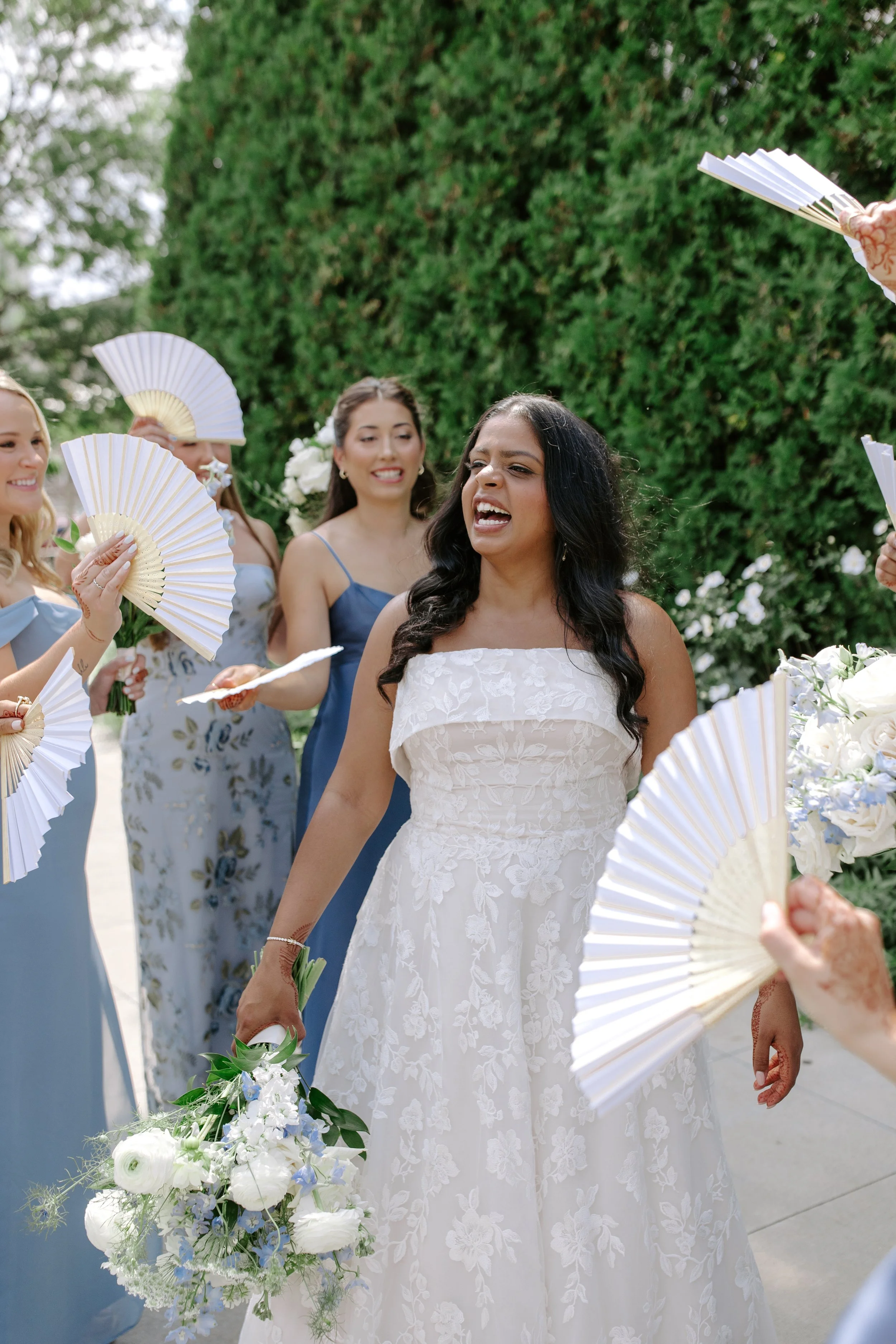 A bride in a white wedding dress with floral embroidery stands among women holding white hand fans and bouquets of flowers outdoors with green trees in the background.