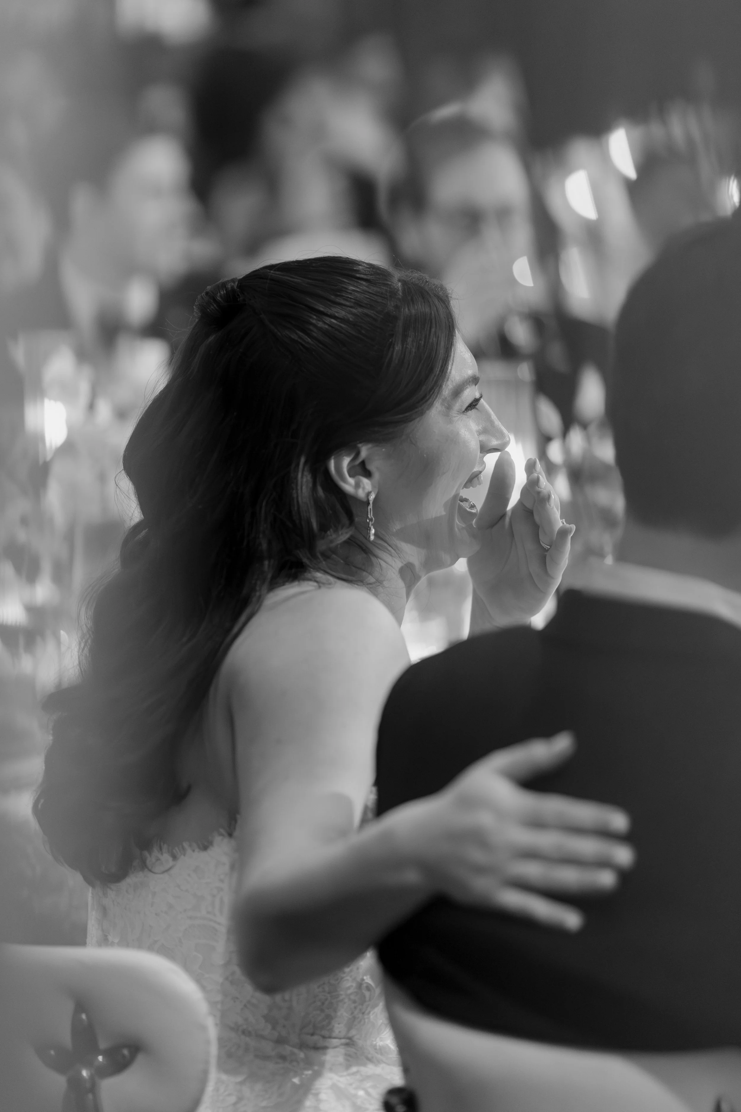 A woman with dark, wavy hair smiling and enjoying a conversation at a formal event, surrounded by other guests in a dimly lit setting.
