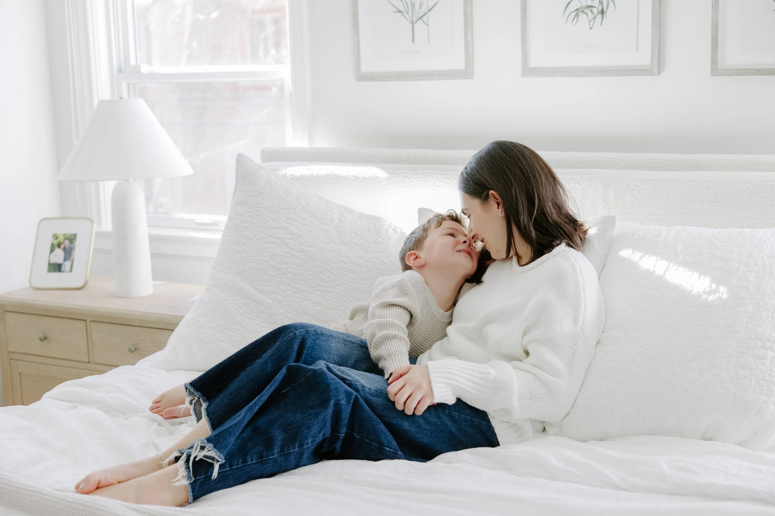 A woman and a young boy lying on a bed sharing a tender moment in a bright, decorated bedroom with framed botanical art on the wall and a side table with a lamp and framed photograph.