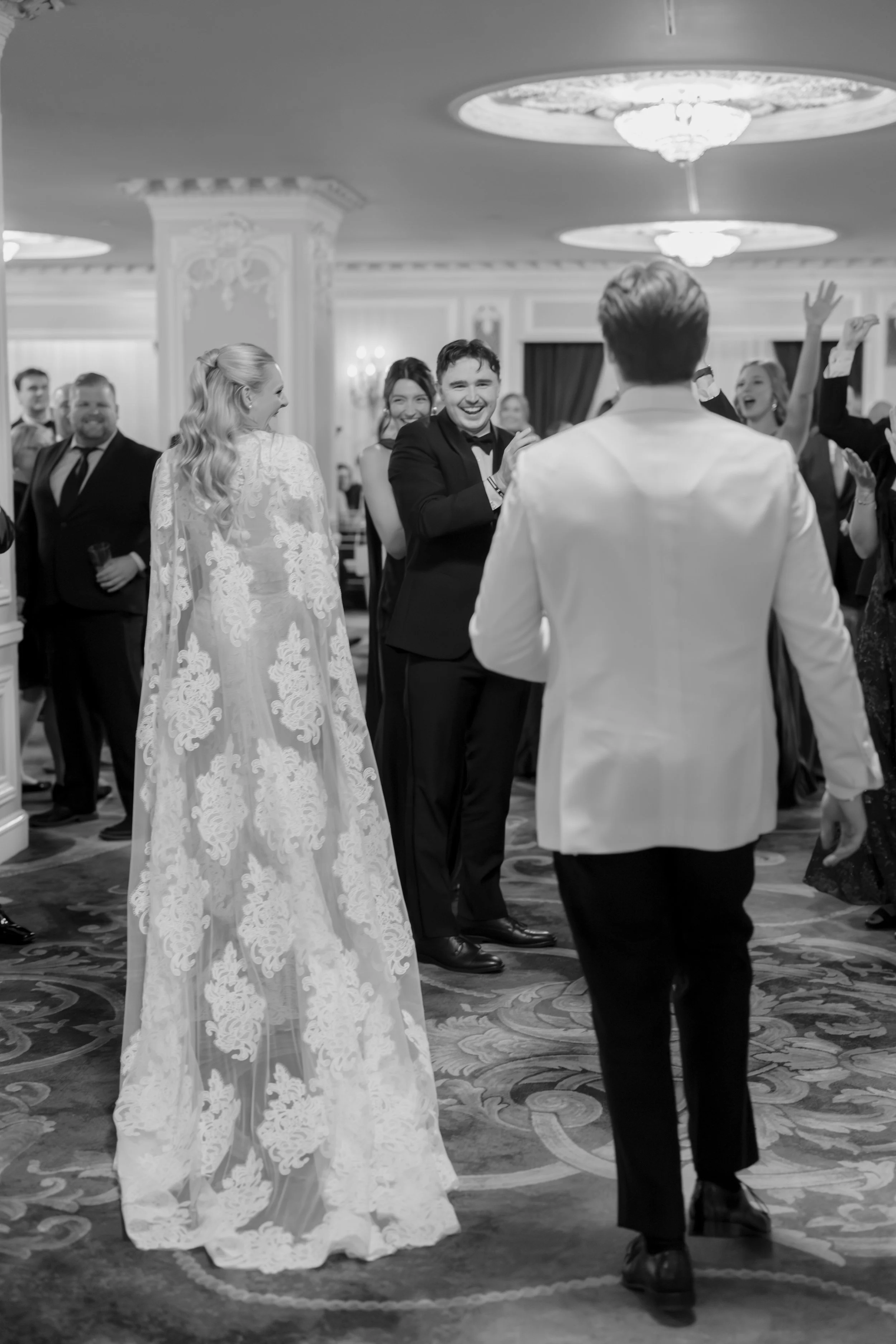 Couple dancing at a wedding reception with a woman in a lace wedding dress and a man in a black tuxedo, surrounded by smiling guests in a decorated ballroom.