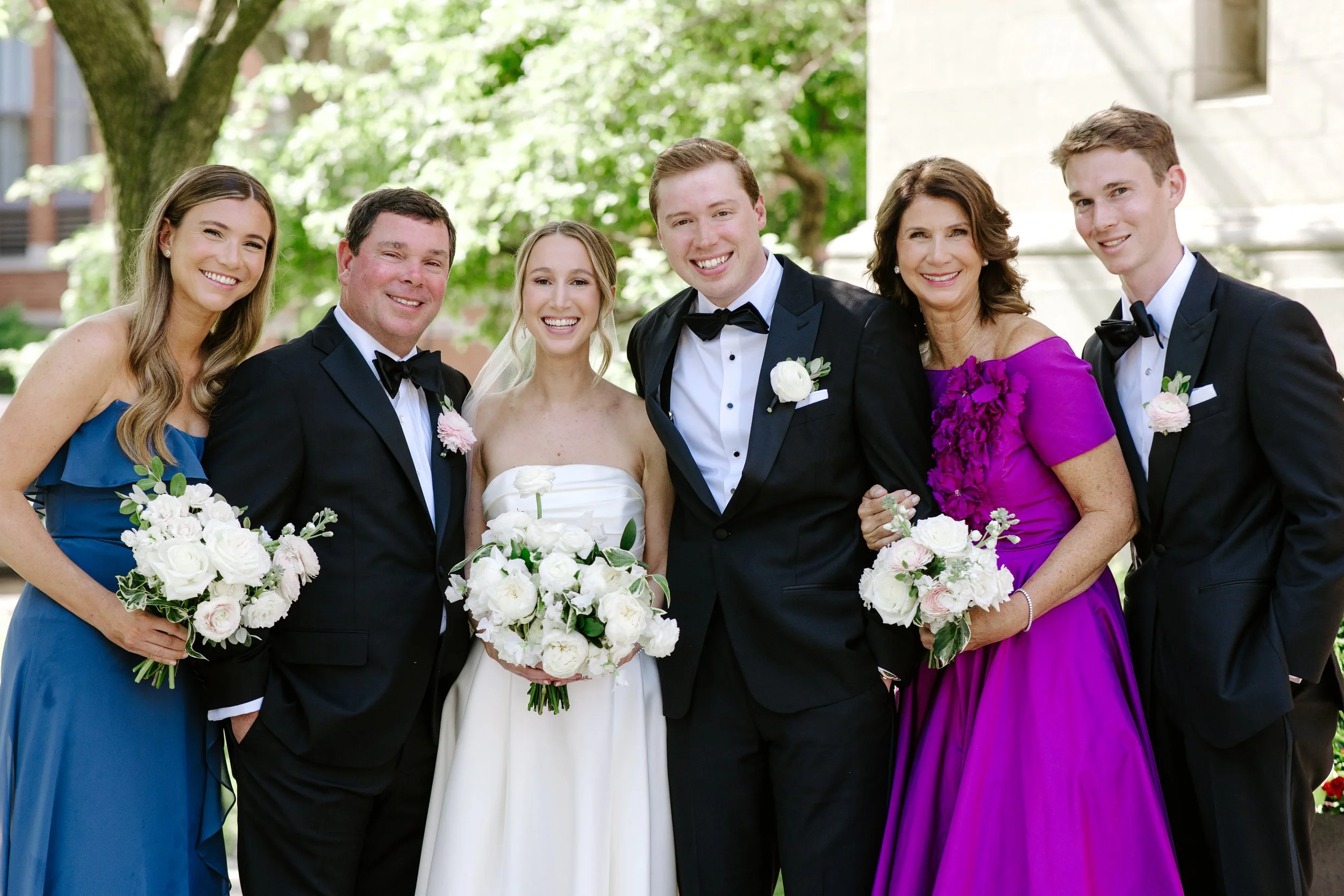 Group of seven people, including a bride and groom, smiling and holding bouquets, at an outdoor wedding with trees and a stone building in the background.