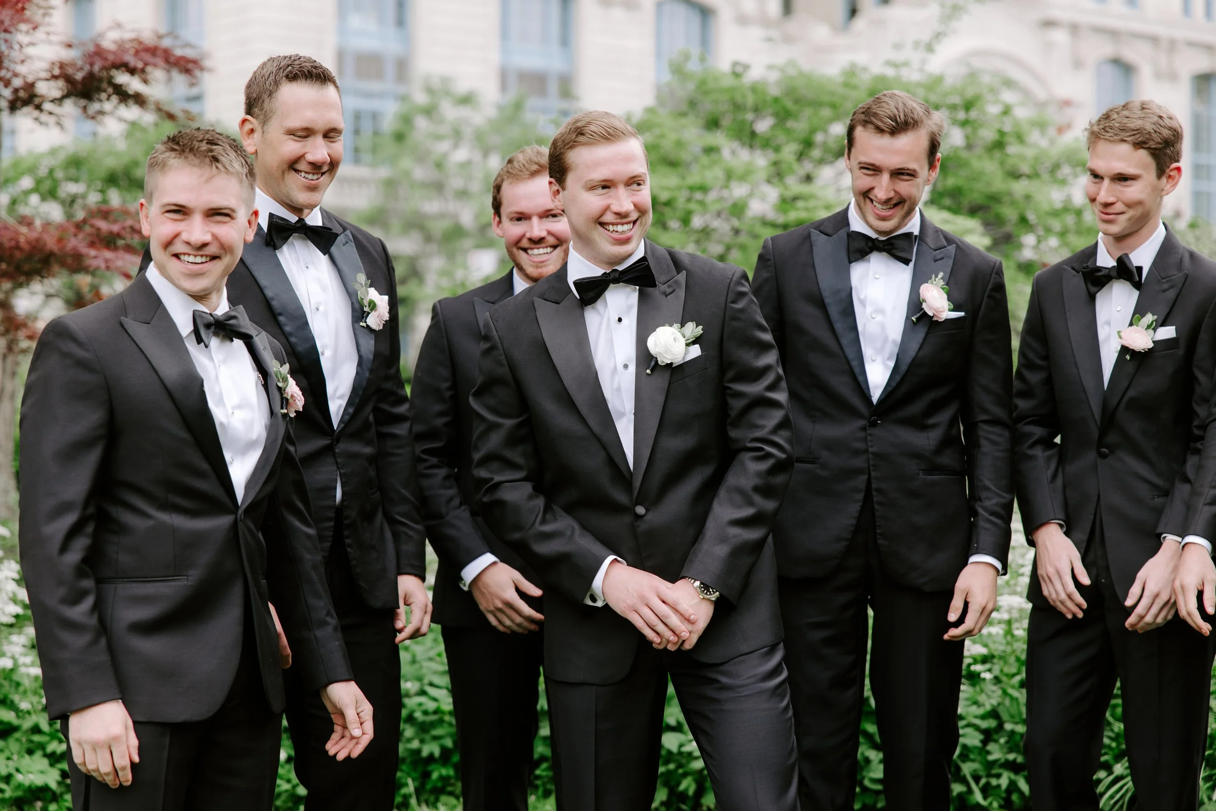 Group of seven men in tuxedos with bow ties and white flowers, standing outdoors in front of a building and green trees, smiling and talking to each other.
