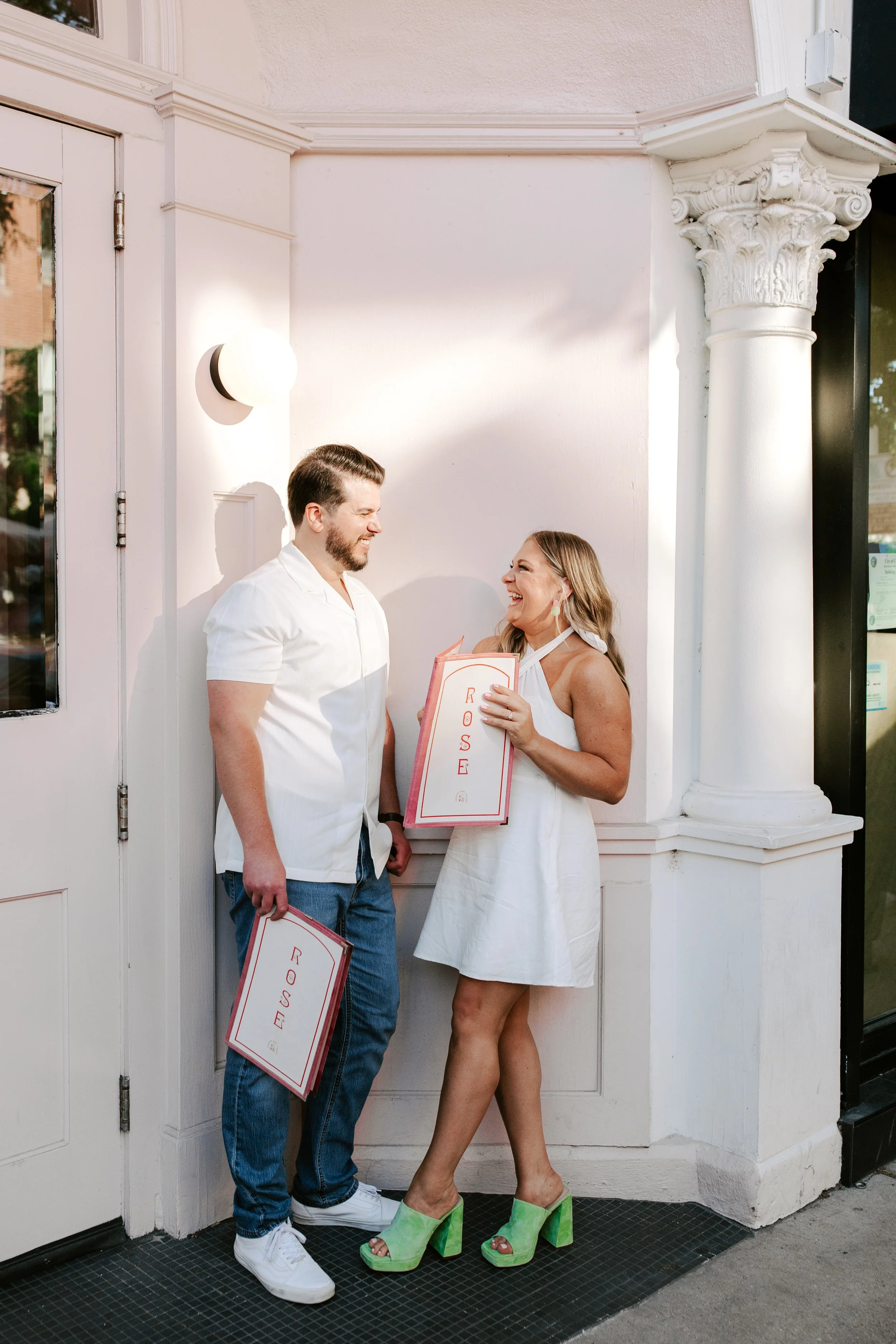 Two people, a man and a woman, standing outside near a white building with decorative columns. They are smiling and holding signs that say 'Rise' and 'Rose'. The woman is wearing bright green high-heeled shoes and a white dress, while the man is in a