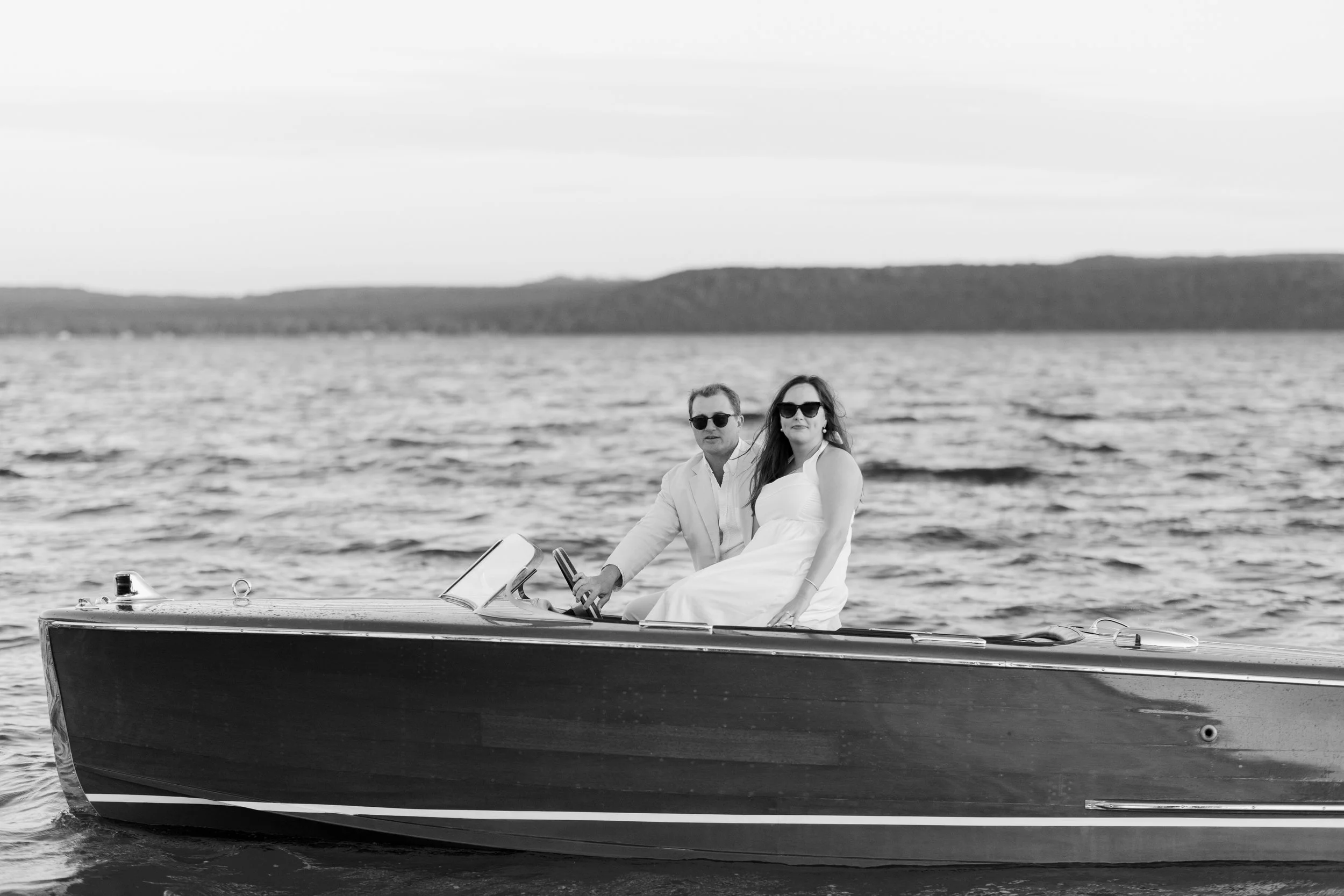 A man and woman are sitting on a boat with the woman wearing a white dress and the man in a light-colored suit, both wearing sunglasses, on a body of water with a distant shoreline in the background.