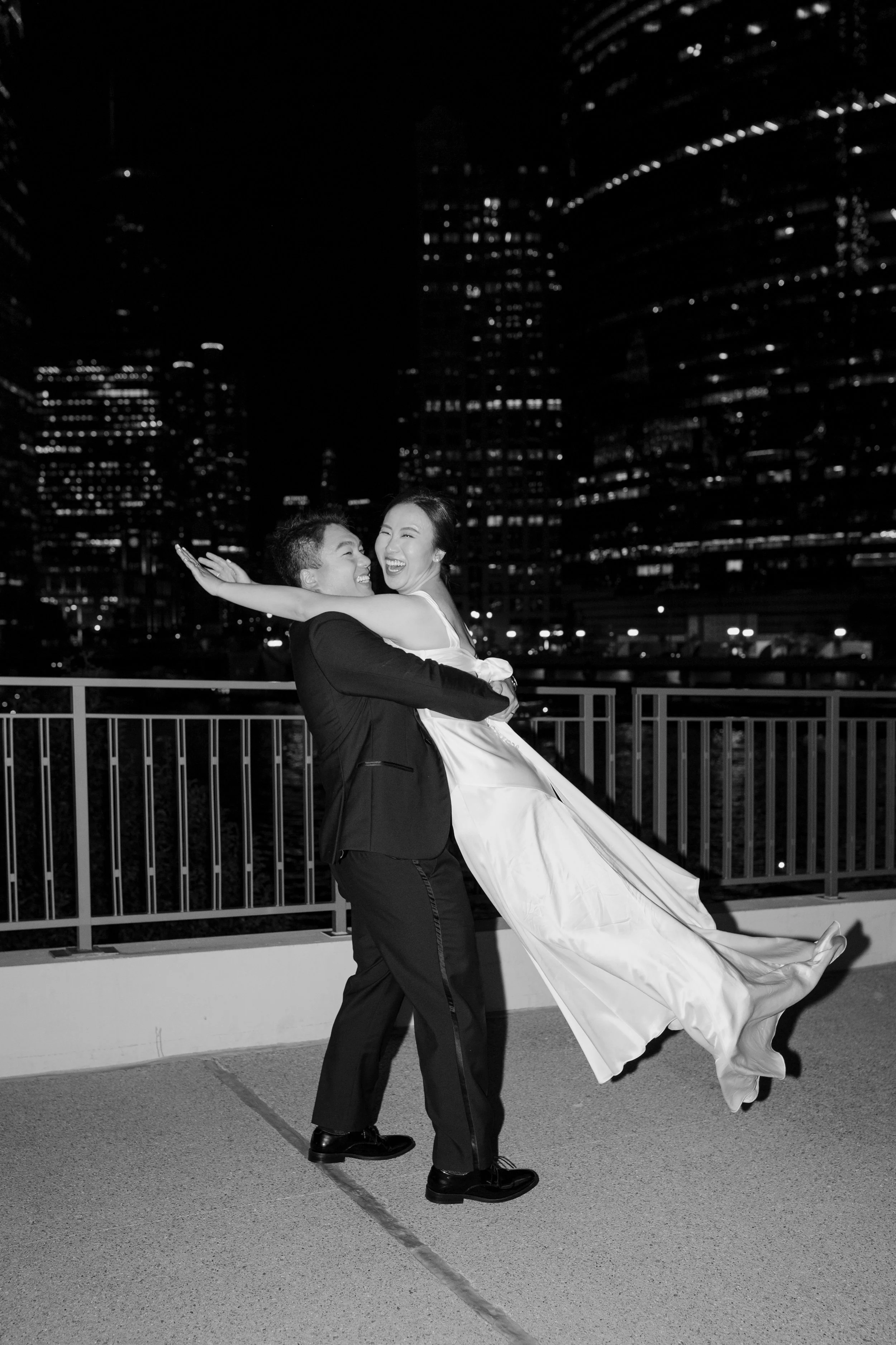 A happy couple dancing on a rooftop at night with city lights and skyscrapers in the background. The man lifting the woman who is wearing a flowing dress.