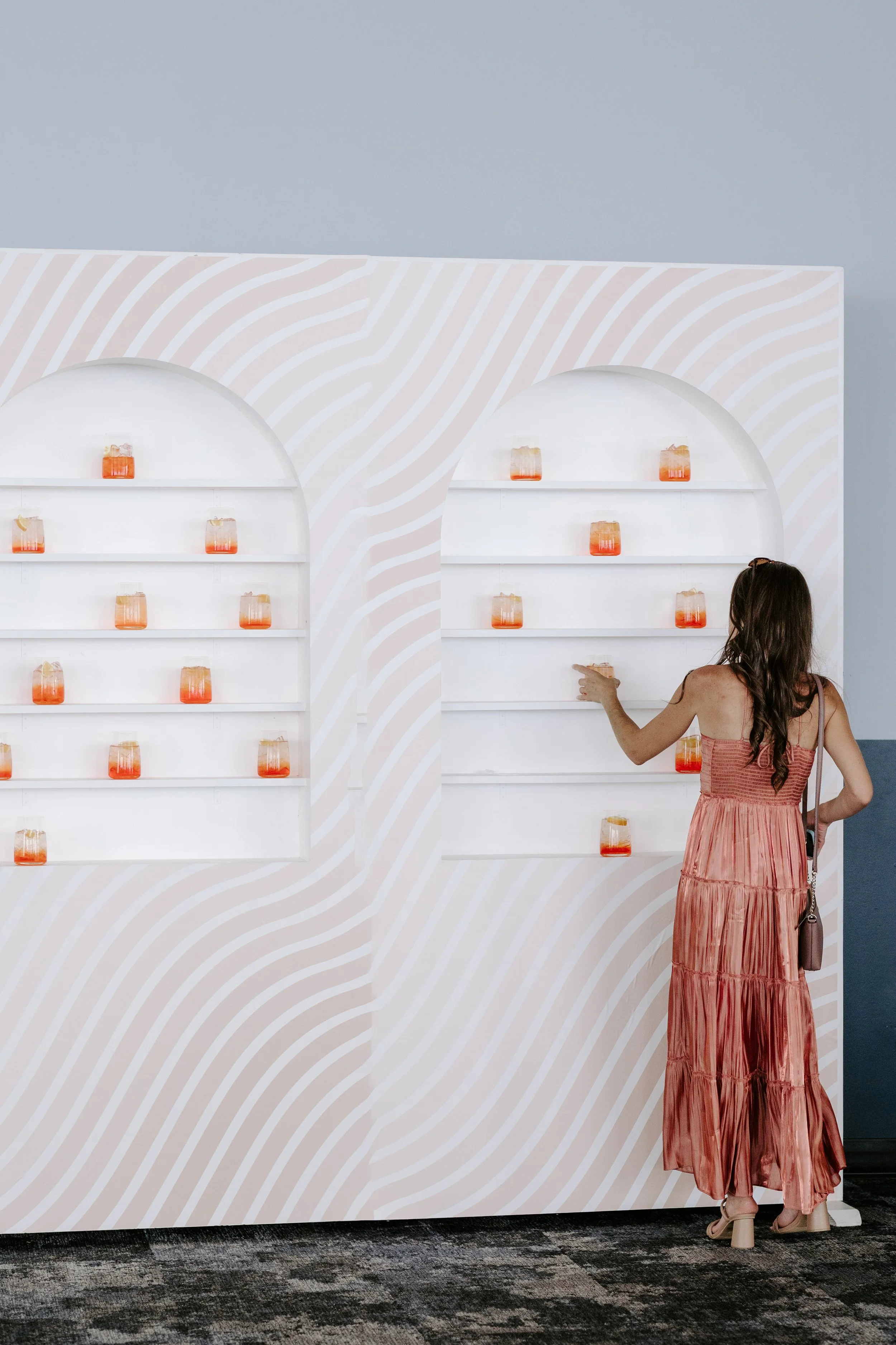 A woman in a pink, pleated maxi dress is standing in front of a white wall with decorative shelves holding orange and pink glass jars.
