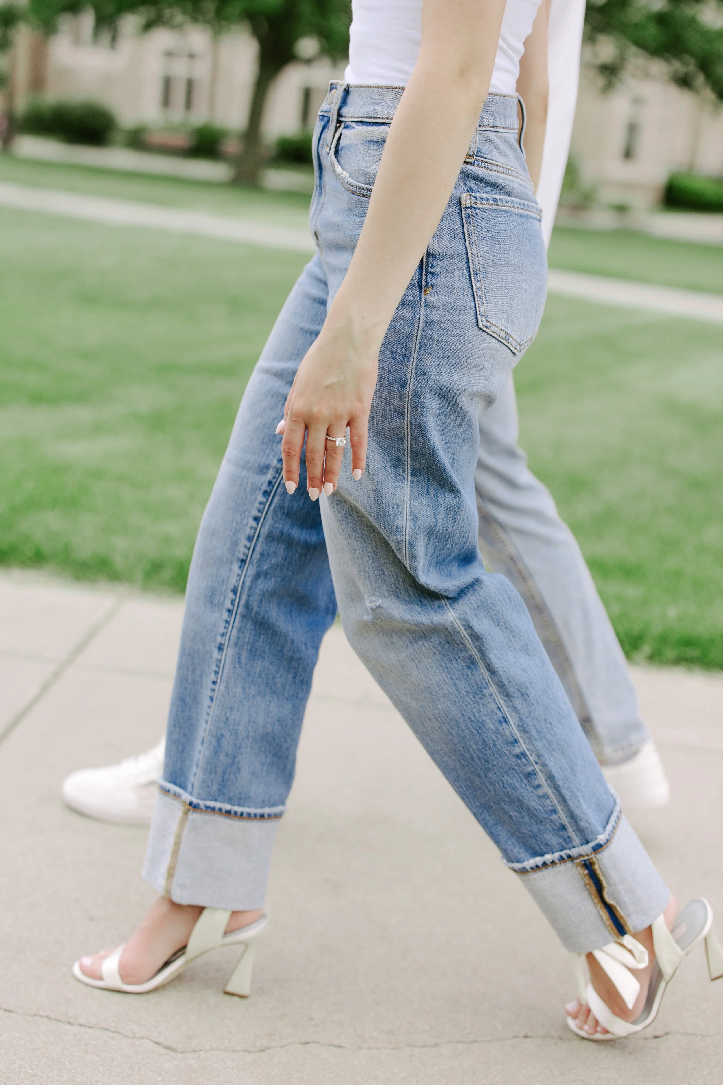 Two women wearing light blue jeans and high heels walking outdoors on a concrete sidewalk with grass and trees in the background.