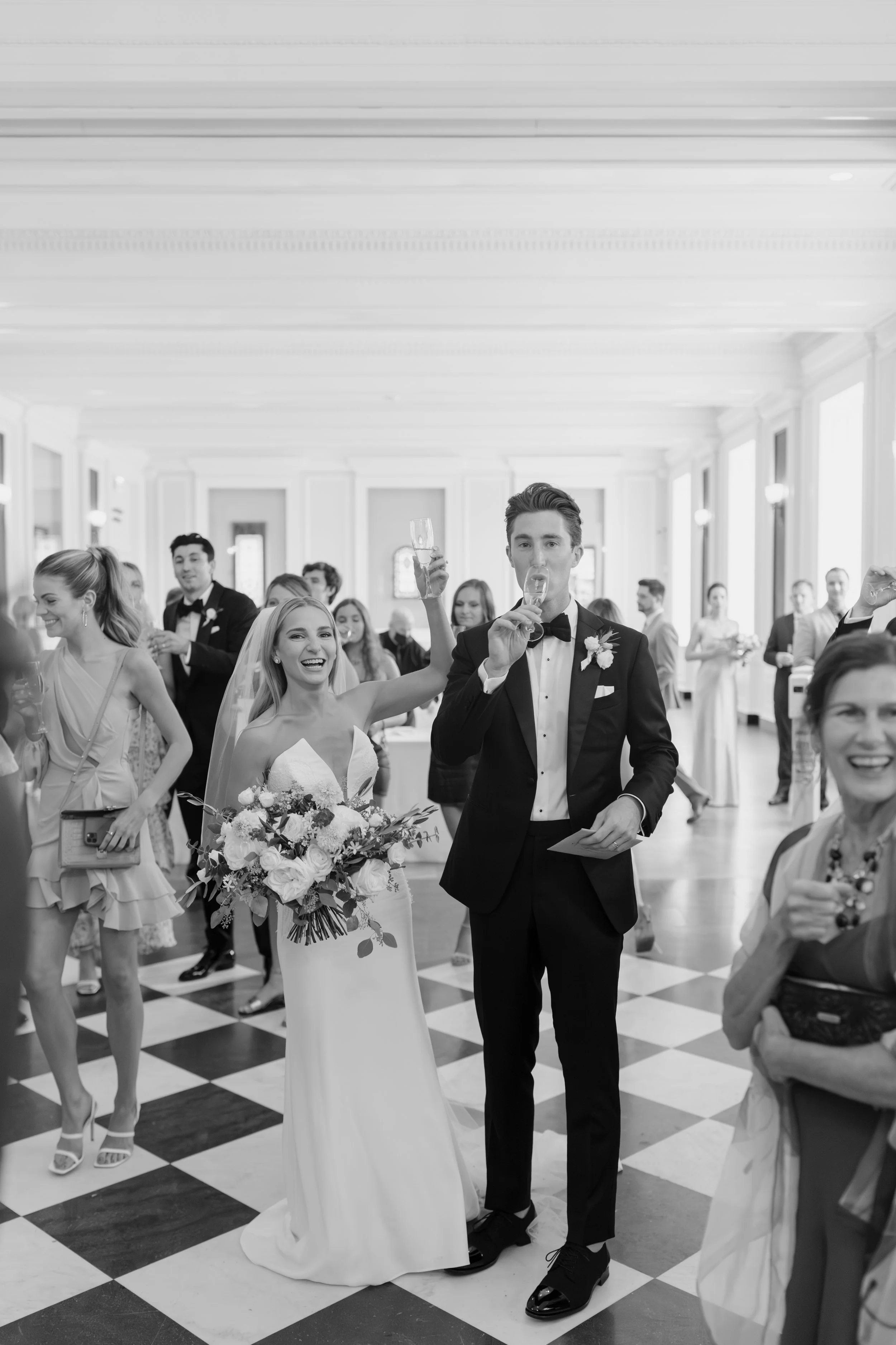 People celebrating at a wedding reception in an elegant, decorated venue with checkered flooring, many guests dressed in formal attire, and a bride holding a large bouquet of flowers, smiling and raising a glass with a groom in a tuxedo.
