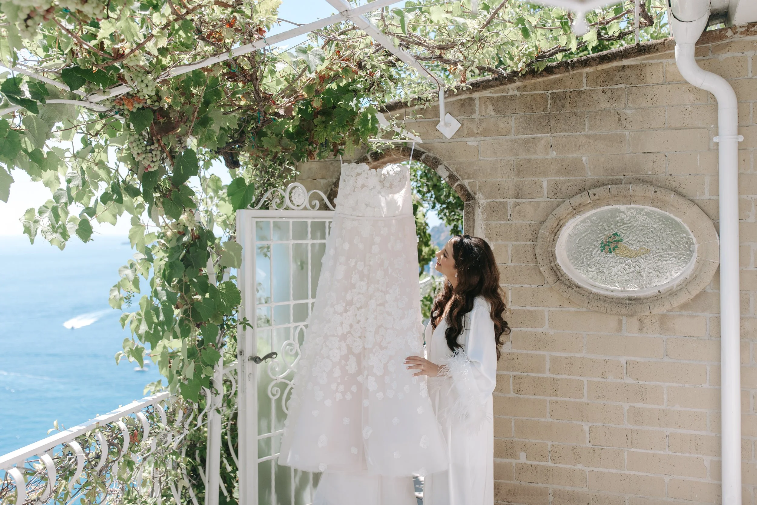 A woman with long, dark hair and wearing a white outfit standing beside a white decorative screen outdoors. The background features a stone wall with a circular decorative element and a water fountain. Green vines with white grapes and leaves grow ov