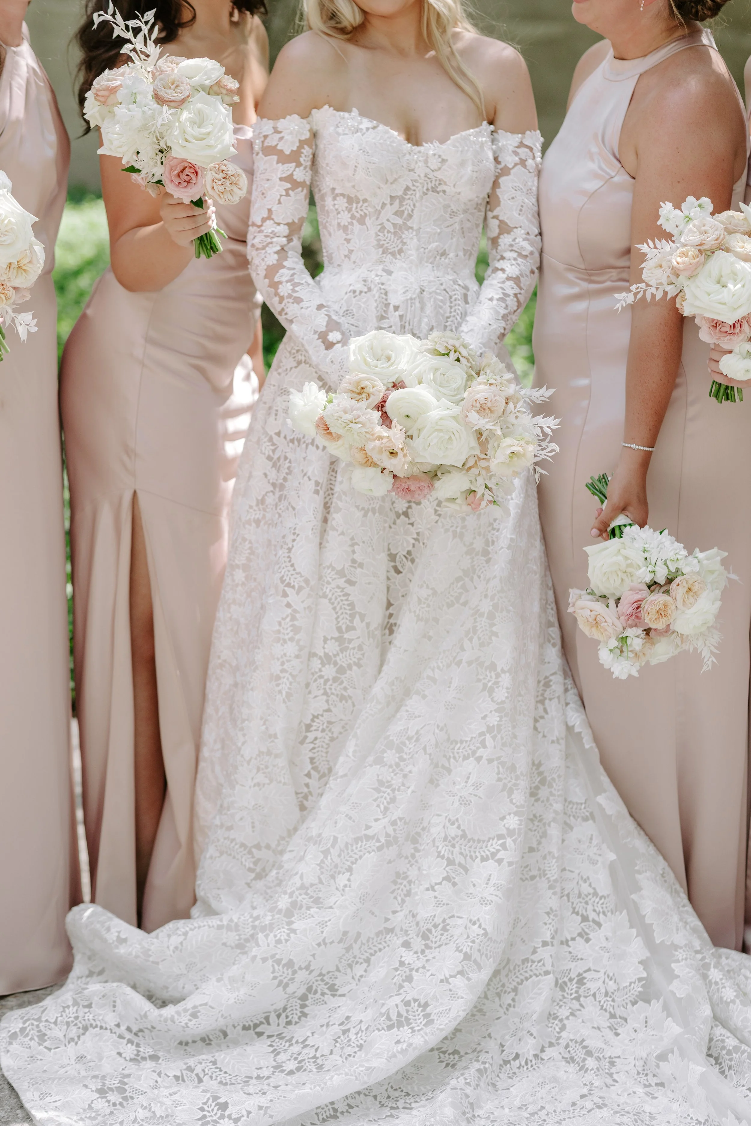 Close-up of a bride in an off-the-shoulder lace wedding dress holding a bouquet of white and blush pink roses, with bridesmaids in blush satin dresses surrounding her, holding similar bouquets.