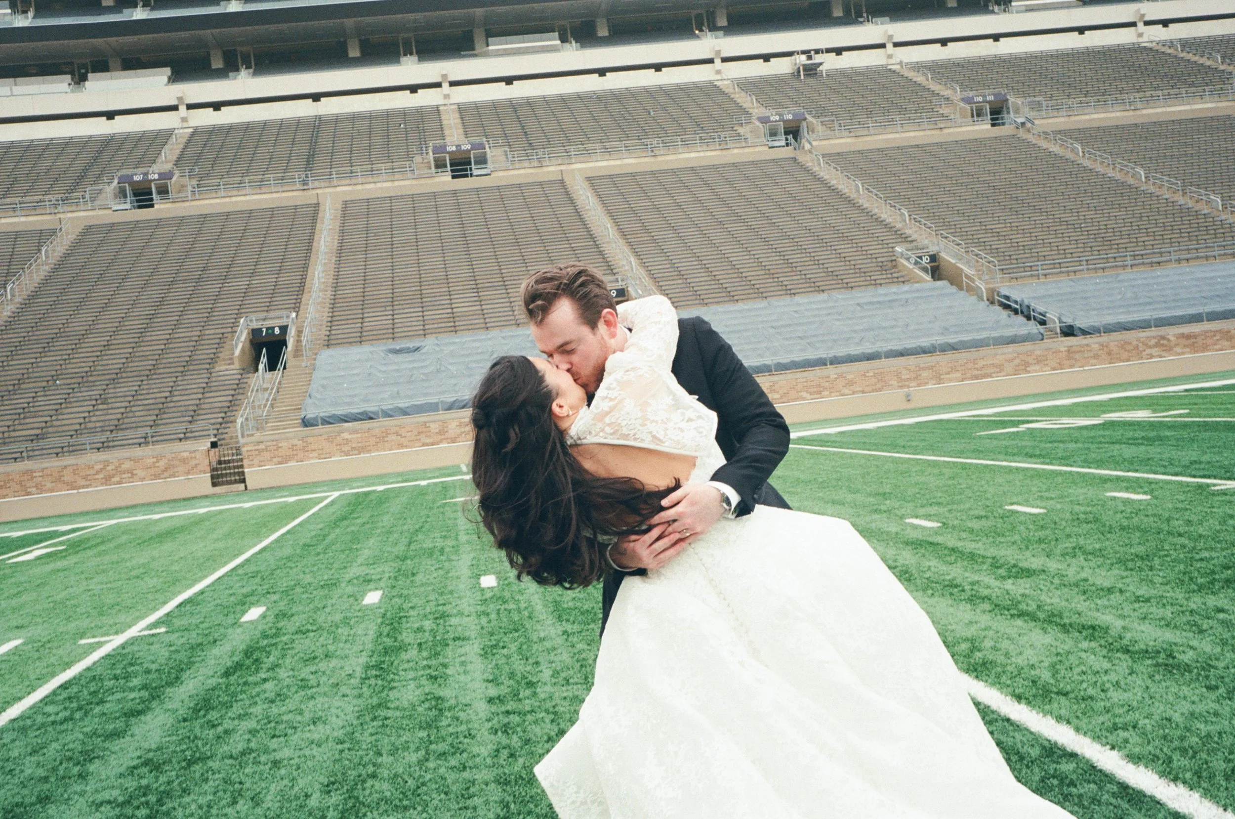 A couple dancing together on the football field of an empty stadium, with the man in a black suit and the woman in a white wedding dress, sharing a kiss.