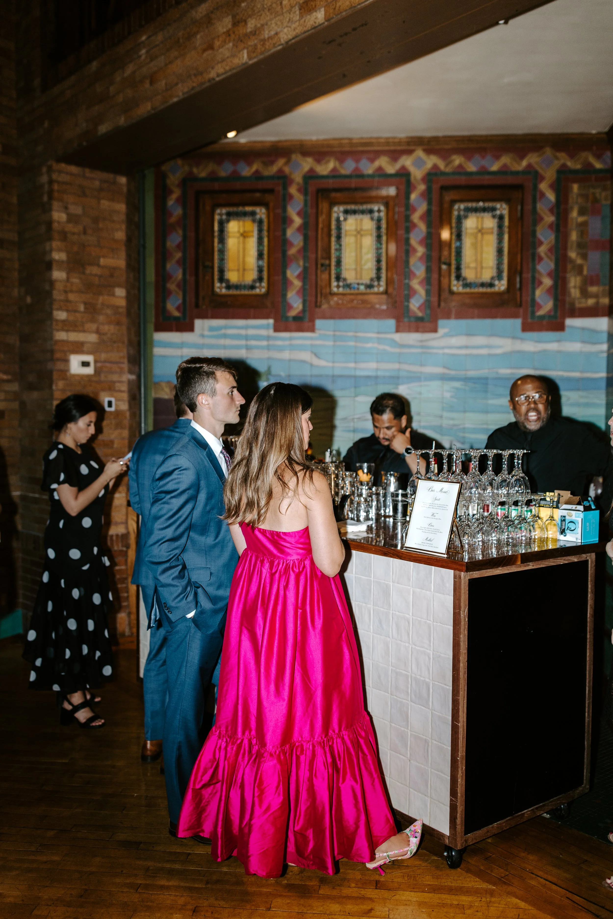 People gather around a bar at a social event, with a woman in a bright pink dress and floral shoes in the foreground, and others in formal attire. The background features a mural and ornate window frames on a brick wall.