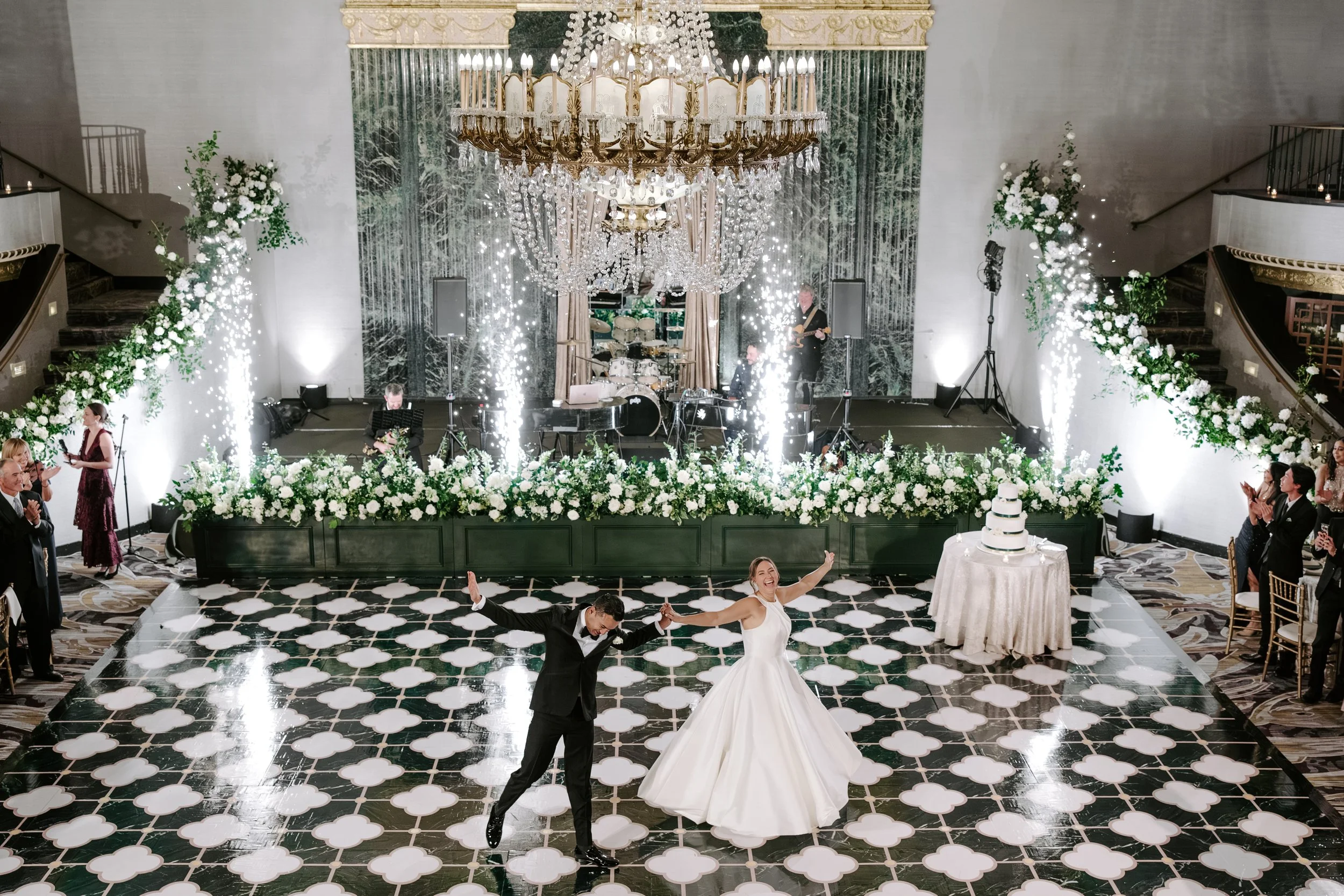 A bride and groom are dancing in the center of a lavish wedding reception hall, surrounded by guests, floral decorations, and a grand chandelier.