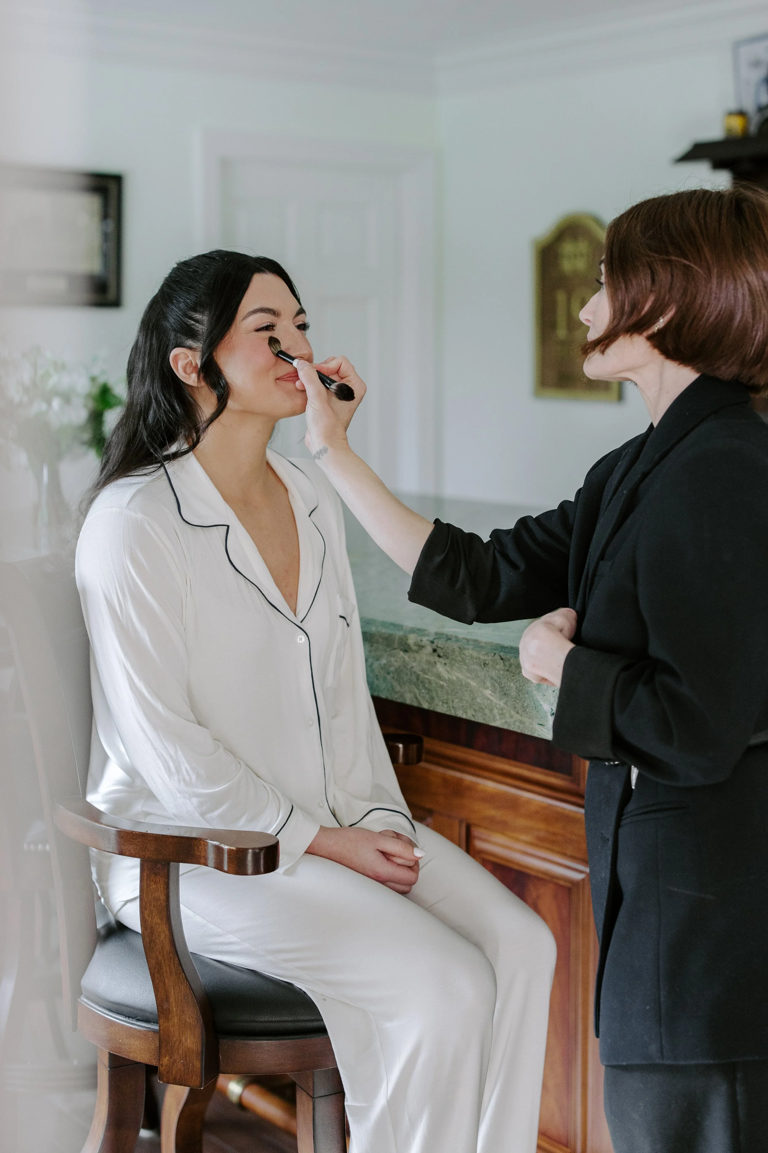 A woman in white pajamas sitting on a wooden chair, having makeup applied to her face by a woman in a black blazer, inside a room with a countertop and a wall with picture frames.
