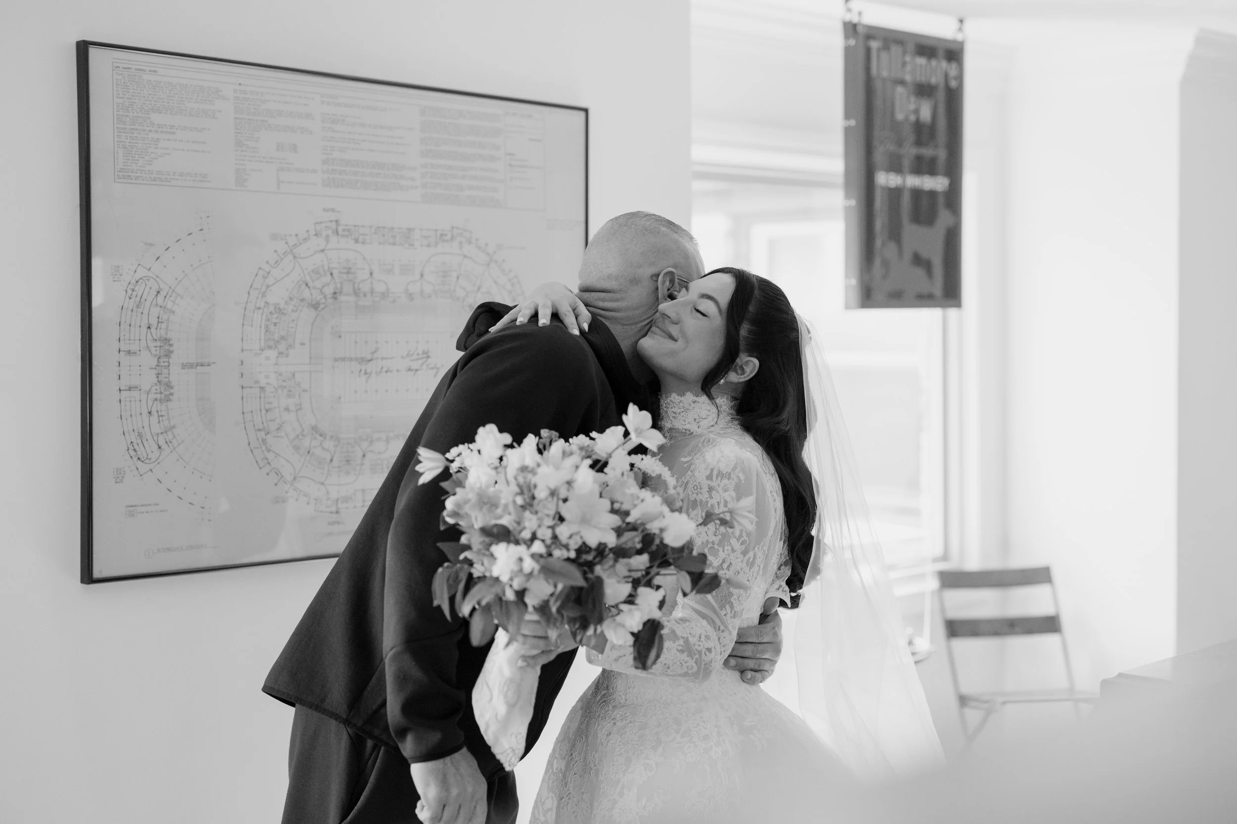 A bride and groom share a tender embrace during their wedding reception, with the bride holding a bouquet of flowers and smiling. The groom's face is hidden as he leans into her. They are standing near a wall with architectural drawings and a framed 