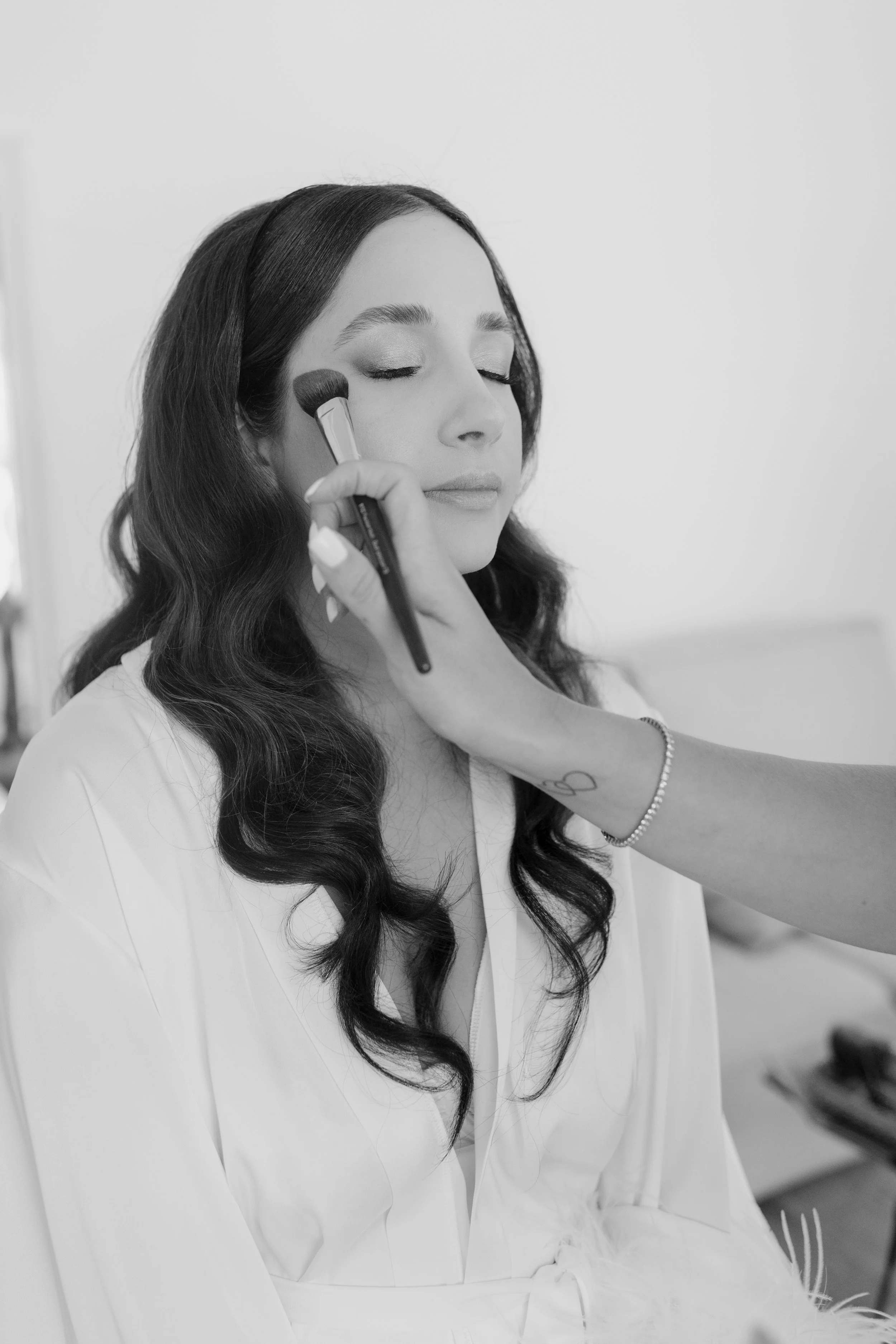 A woman with long dark hair, eyes closed, applying makeup with a brush on her cheek, wearing a white outfit and a bracelet, in a bright room.