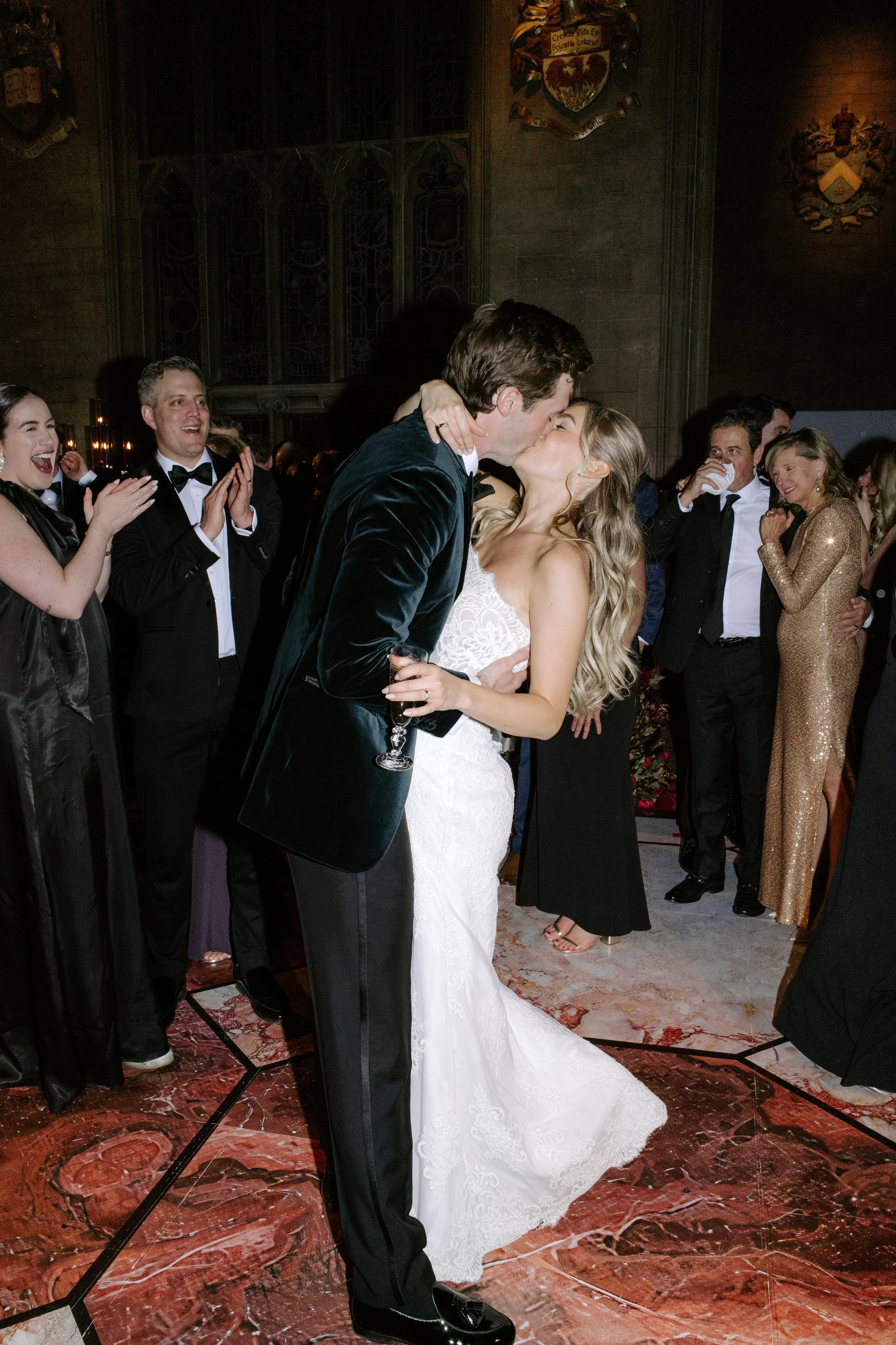 A couple in wedding attire sharing a kiss at their wedding reception, surrounded by friends and family in an elegant venue with stained glass windows and coats of arms on the walls.