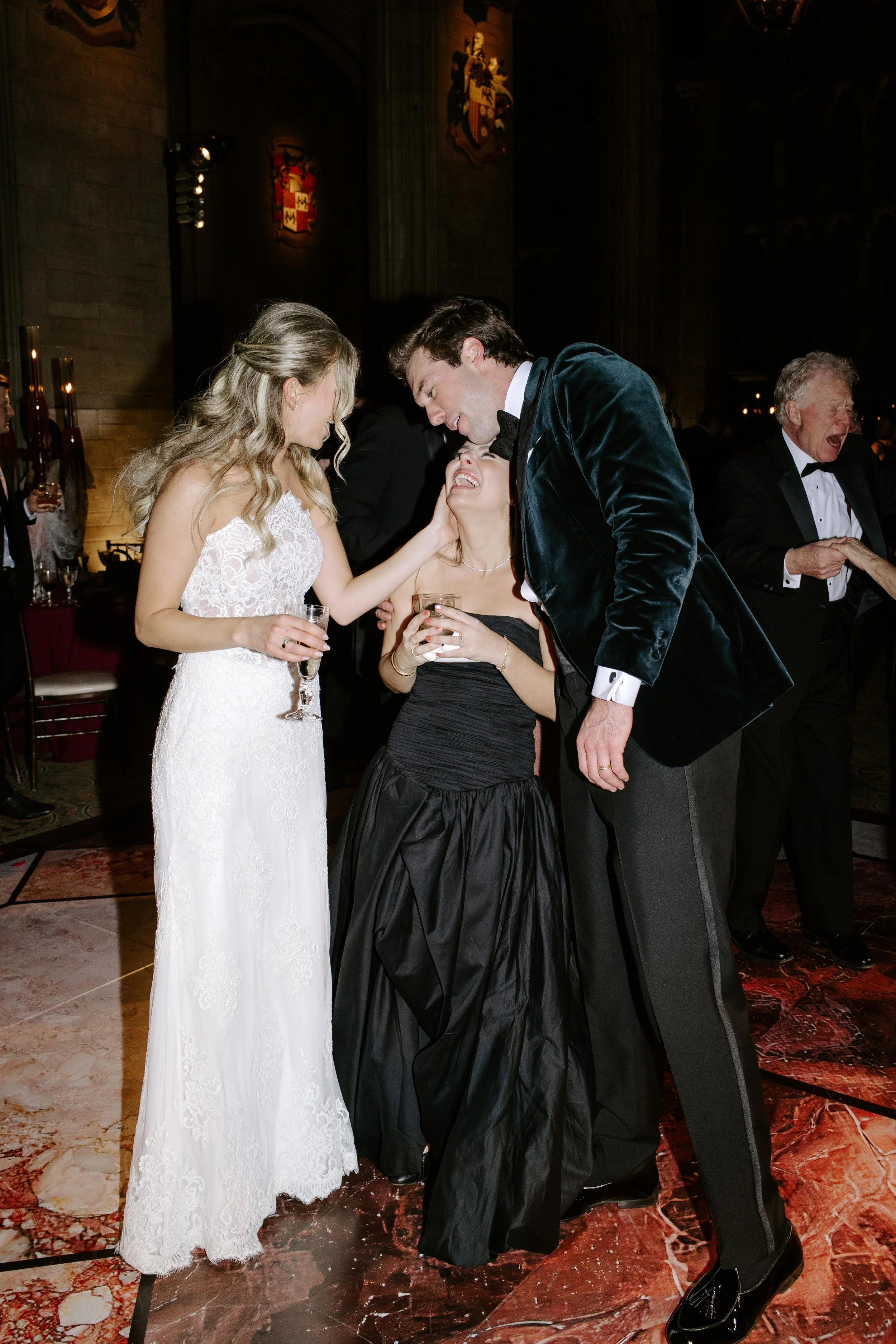 A wedding reception with a bride in a white lace gown and two men, one in a tuxedo and the other in a velvet tuxedo jacket, smiling and interacting with a woman in a black dress. In the background, an older man is singing or speaking, and a coat of a