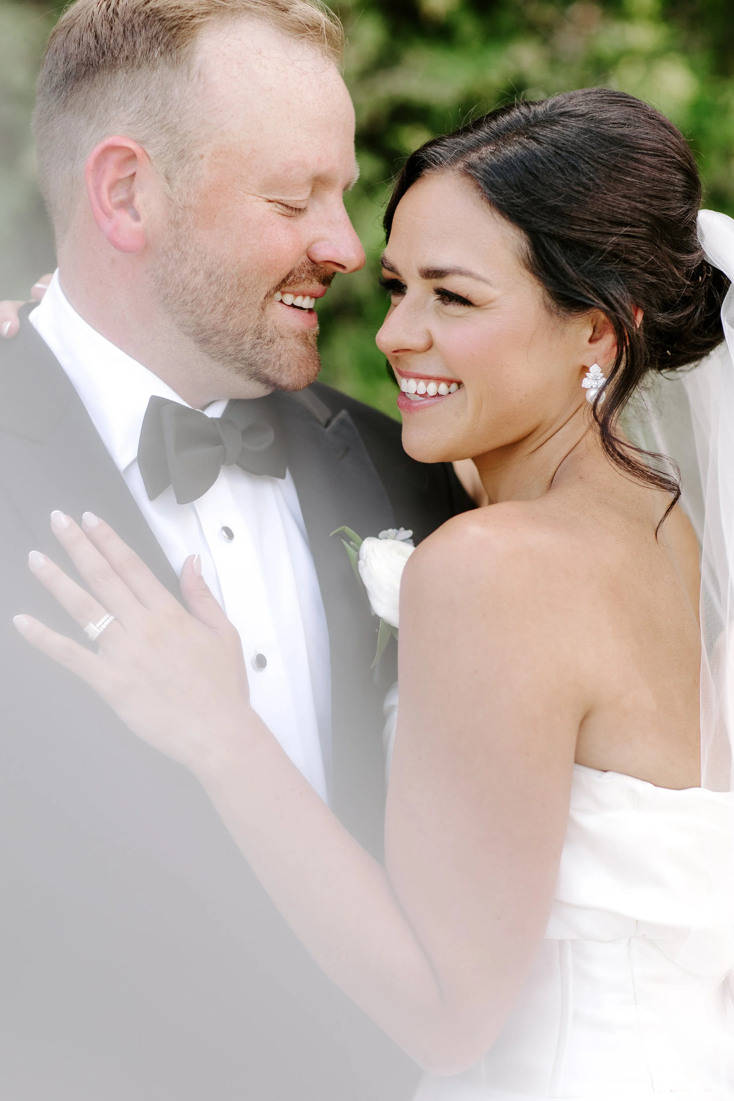 A bride and groom smiling close together, the bride wearing a white dress and the groom in a tuxedo with a bow tie, outdoors with greenery in the background.
