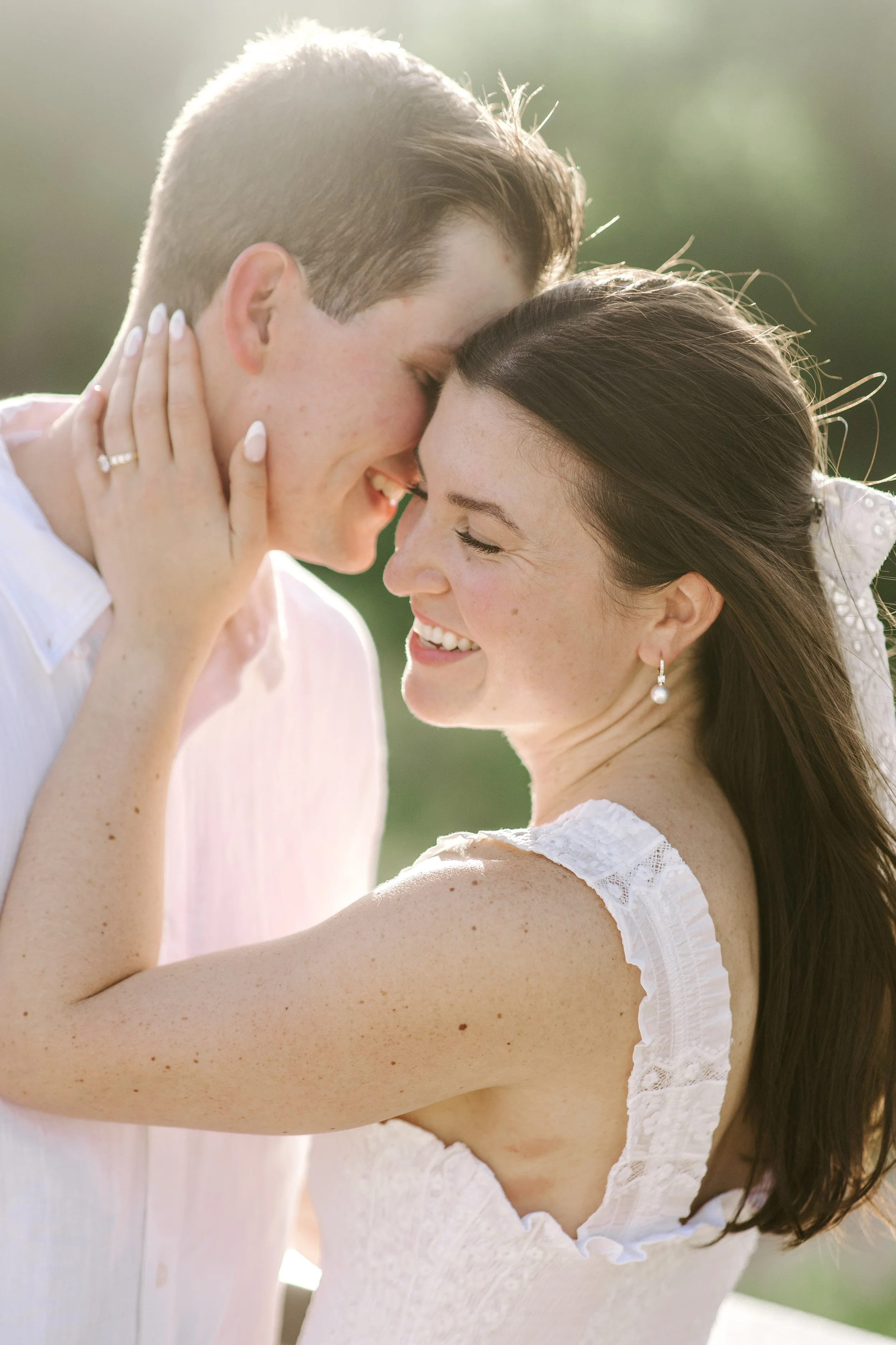 A couple smiling and touching foreheads, outdoors during daylight, with the woman wearing pearl earrings and a white dress.
