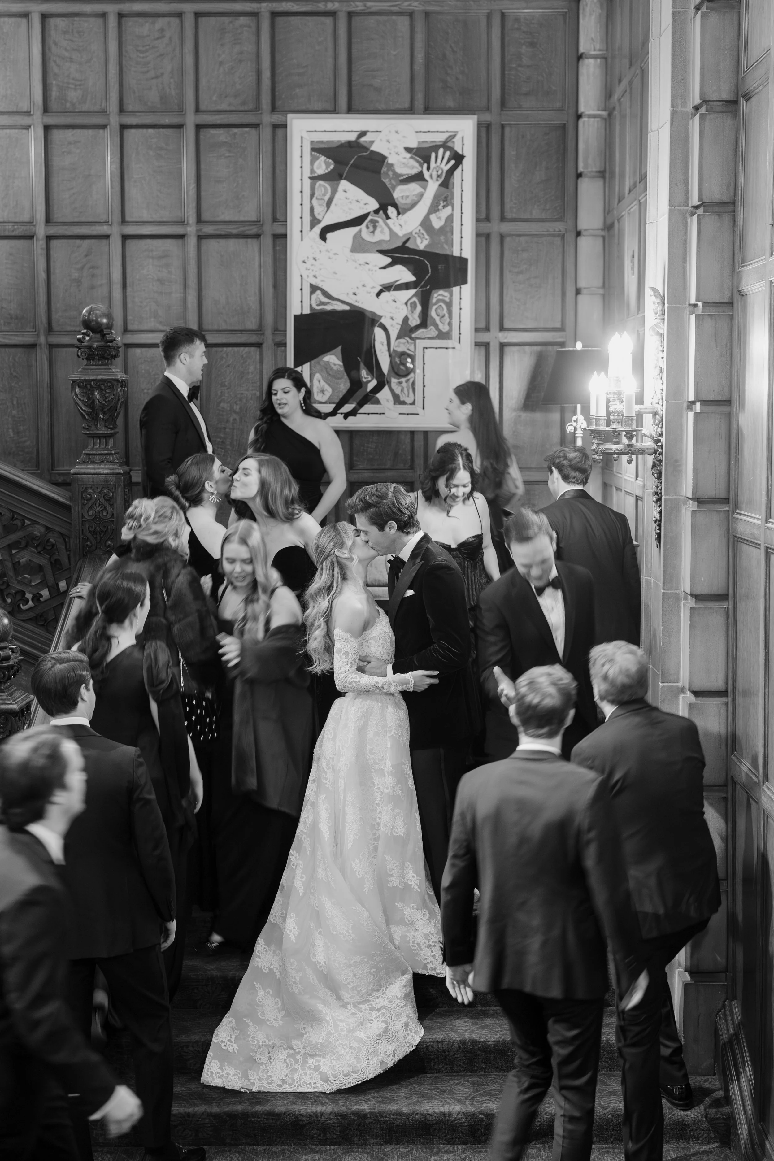 A black and white photo of a wedding reception with a bride and groom dancing on a staircase surrounded by guests in formal attire. The setting has wood-paneled walls, a large abstract painting, and a candelabra on the wall.