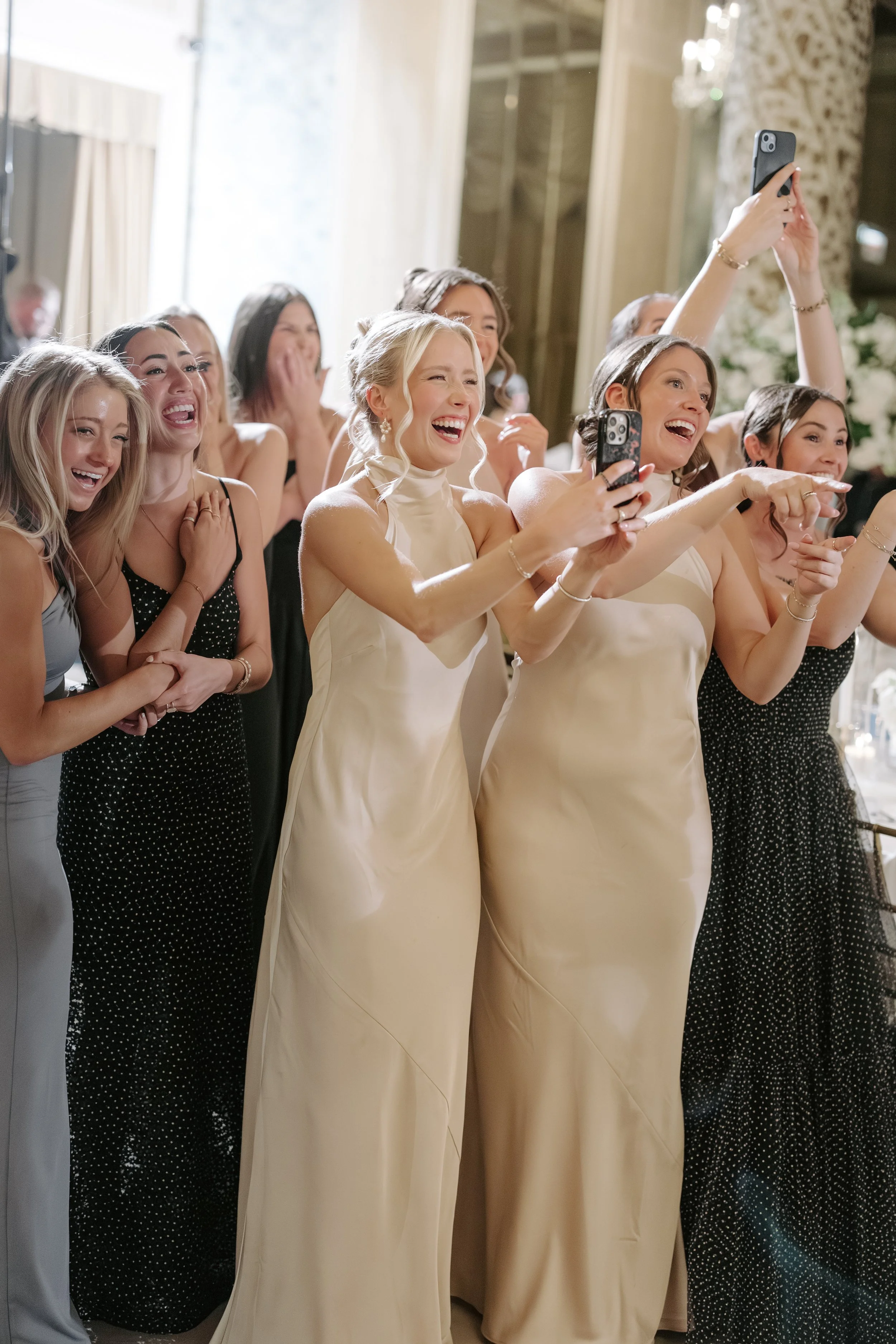 Group of women laughing and taking selfies at a celebration, dressed in formal attire, indoors with elegant decor.