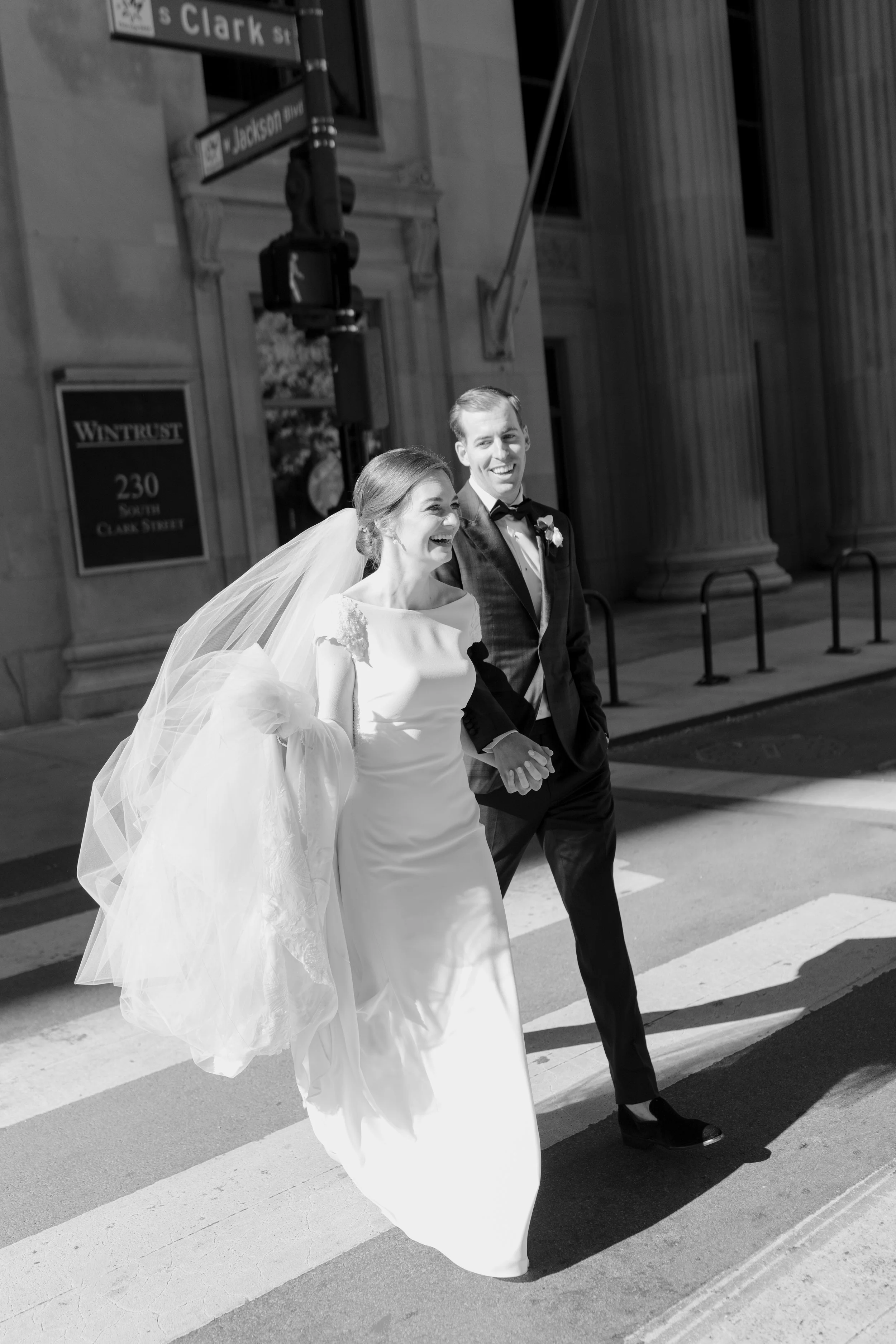 A bride in a white wedding dress and a groom in a black tuxedo holding hands and smiling while walking on a city street at night.