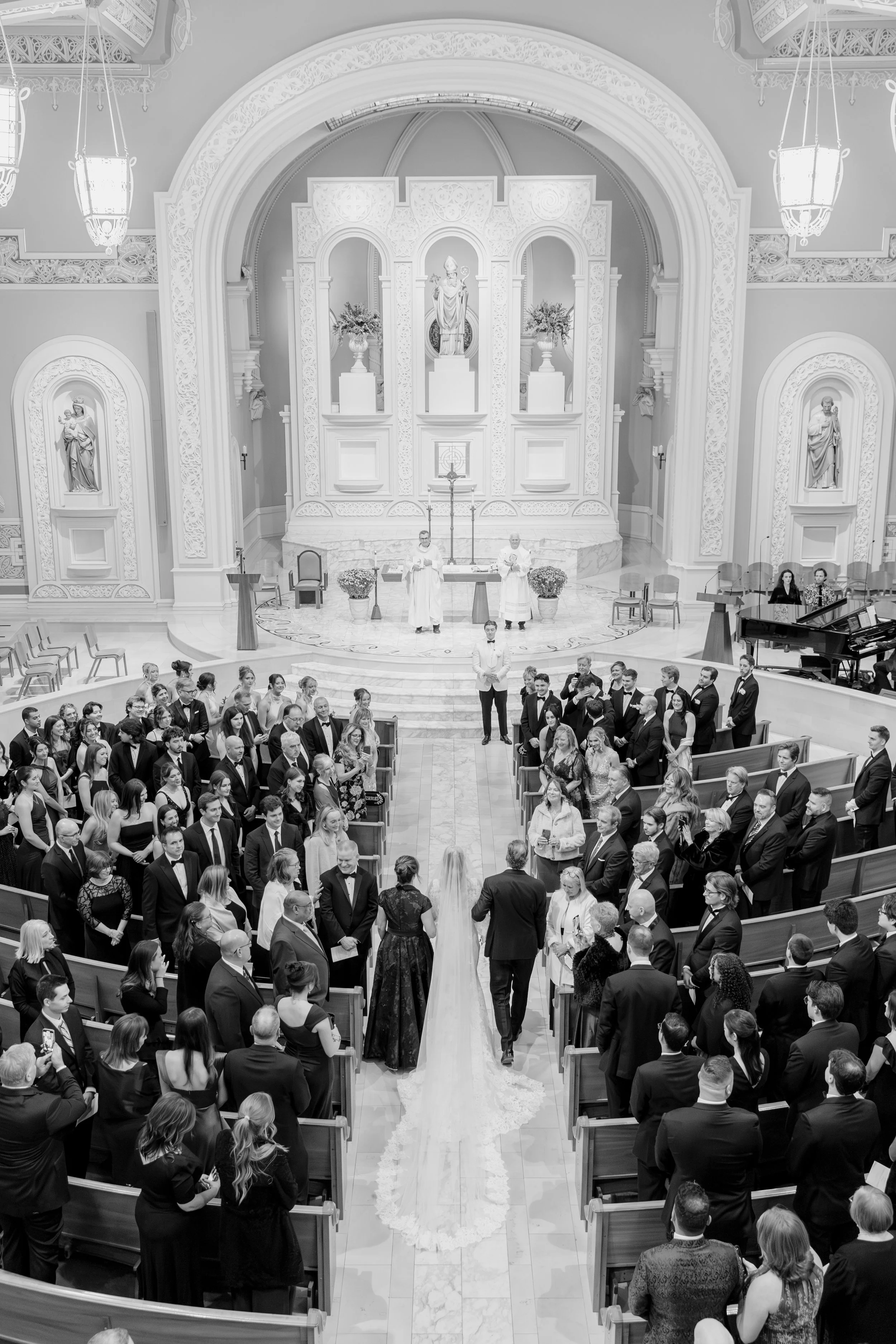 A wedding ceremony inside a church, with a bride in a white gown and a groom in a tuxedo standing at the altar. The priest and wedding officiants are behind the couple, and wedding guests are seated in pews, watching them.